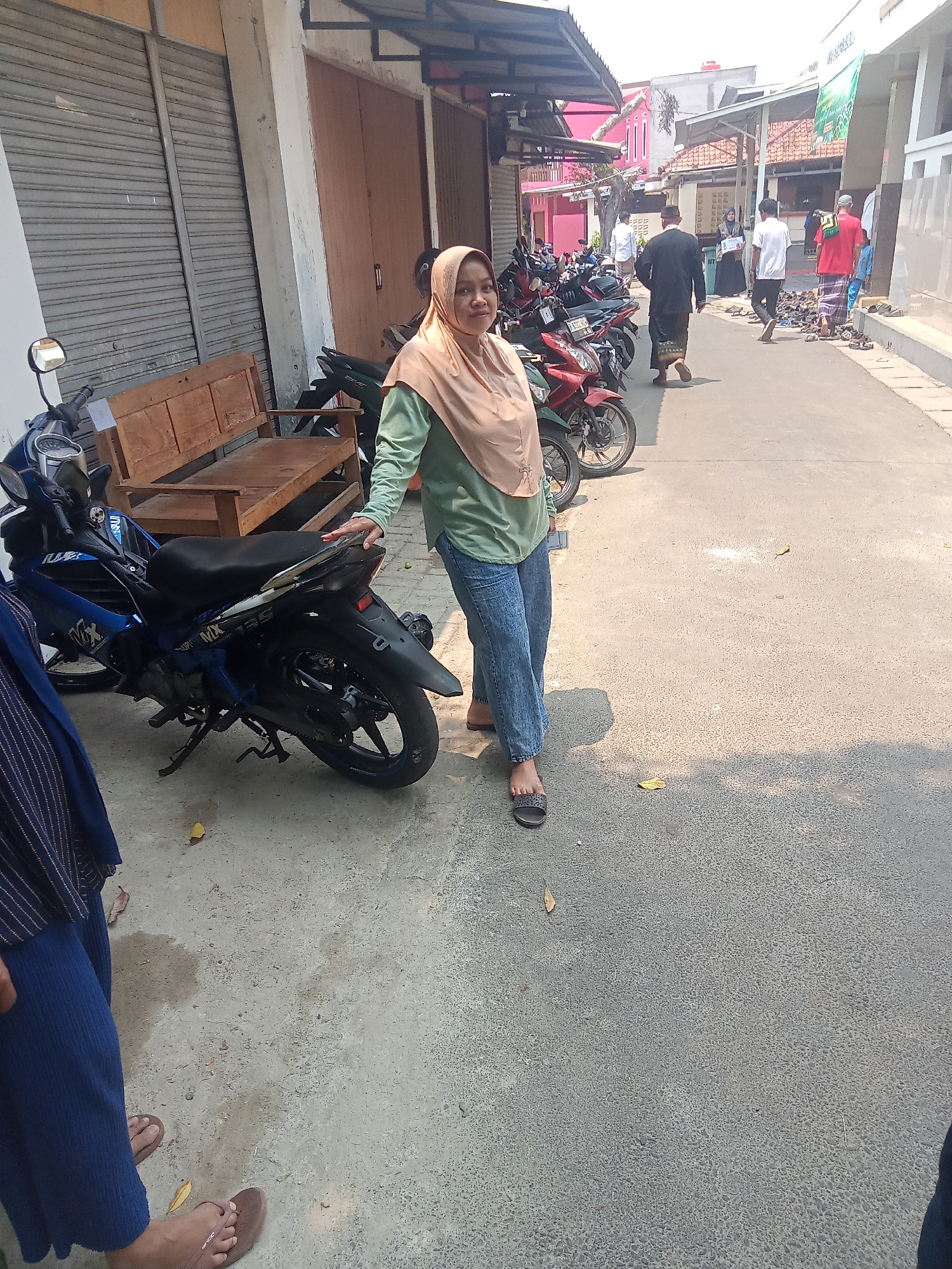 The image depicts a street scene in the city of South Tangerang, Indonesia, likely in a residential or commercial area with shops. A woman wearing a light brown hijab and a green long-sleeved shirt is standing next to a blue motorcycle, with her hand resting on its seat. She is wearing blue jeans and dark sandals. To the left of the woman, another person is partially visible, wearing a dark blue garment. Behind the woman, a wooden bench is situated in front of a building with closed gray roller shutters. Further down the street, several other motorcycles are parked. In the background, more buildings are visible, along with a few people walking, some of whom are wearing traditional Indonesian attire. The ground is paved with asphalt, and the scene is illuminated by natural daylight.