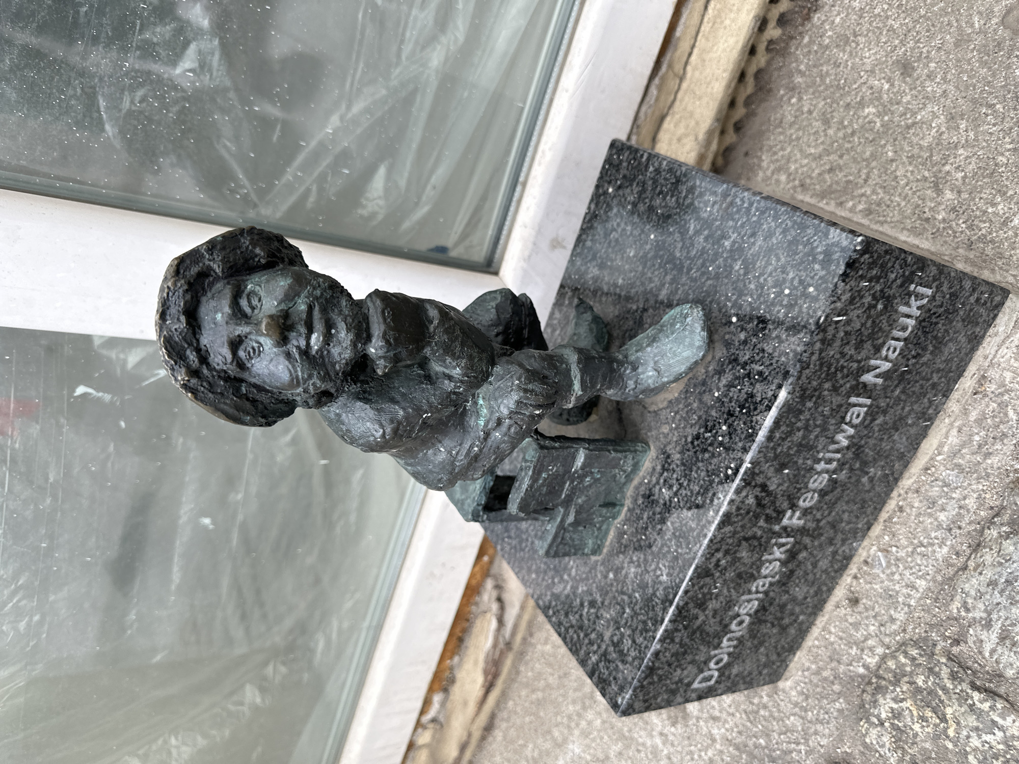 A bronze statue of a gnome sits on a black marble base, labeled "Dolnośląski Festiwal Nauki," indicating it is located in Wrocław, Poland. The gnome is depicted sitting on a small structure, possibly a box or crate. The statue is positioned near a window with a white frame. The ground surrounding the statue is a rough, grey concrete surface.