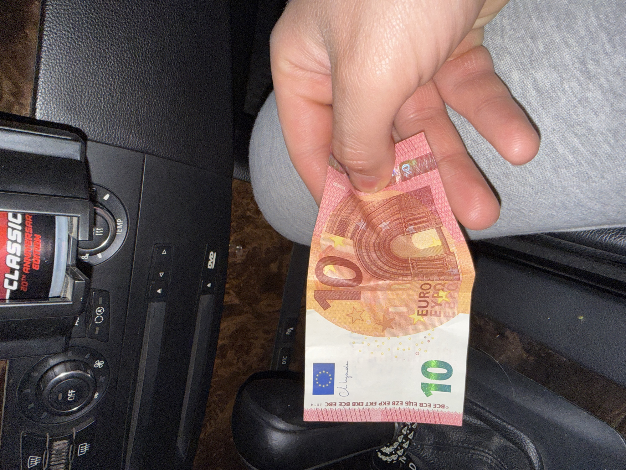 A hand holds a 10 Euro banknote in the interior of a vehicle. The banknote displays the denomination "10" and the Euro symbol. The vehicle's interior includes a climate control panel with temperature adjustments, a DVD player slot, and a CD player with a CD labeled "CLASSIC 20TH ANNIVERSARY EDITION" visible. The hand is positioned over what appears to be a car seat and possibly part of a shoe. The location is unknown.