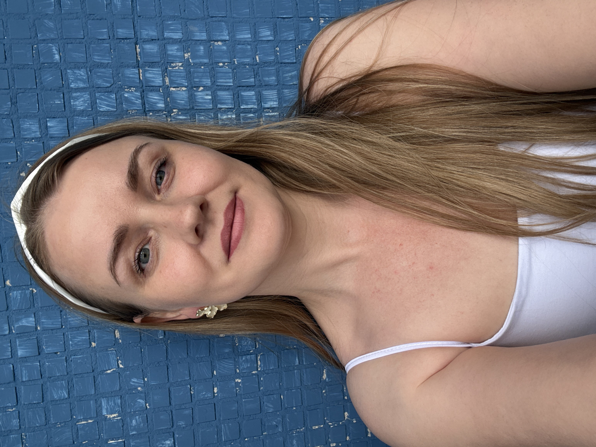 A blonde woman wearing a white tank top and a white headband poses in front of a blue tiled wall. She has long blonde hair and is wearing a flower-shaped earring on her left ear. The image appears to be a selfie, taken outdoors. There is no indication of the city of Chaikovsky, Russia within the image or its context.
