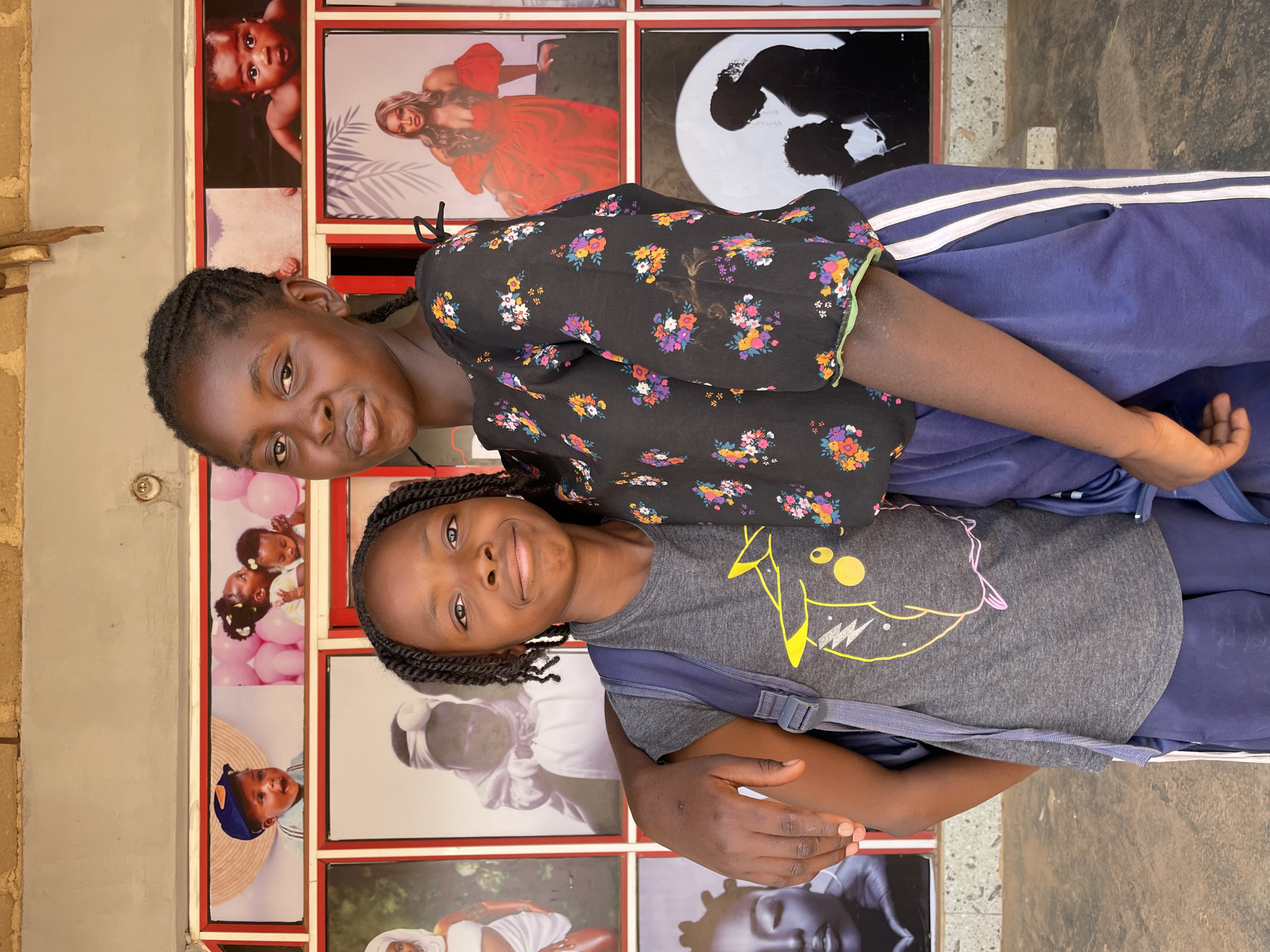 Two girls are posing for a photograph, looking directly at the camera with warm smiles. They appear to be outdoors or in a semi-open public space in Barakin Akawo, Nigeria, standing against a wall covered with a grid of framed portrait photographs.
The girl on the left, who appears older, wears a black top with colorful floral patterns (pinks, oranges, purples, yellows, greens) and dark blue athletic pants with two white stripes running down the side. Her hair is styled in dark braids. The younger girl on the right wears a grey t-shirt featuring a stylized yellow and pink graphic resembling a Pikachu face, along with dark blue pants and has backpack straps visible over her shoulders. Her hair is also in dark braids.
The background is a display of various portrait photographs, possibly from a photography studio or a vendor's stall. These photos include: a baby peeking from the top, a woman in a red dress, a silhouetted couple, three babies with pink balloons, a baby in a blue hat, a woman in a white headwrap, and a close-up of a face that might be a sculpture or artistic rendering. The overall impression is one of bright daylight and a lively, possibly commercial, setting. No specific text is clearly legible on the background photos or surroundings, other than the stylized graphic on the younger girl's shirt.