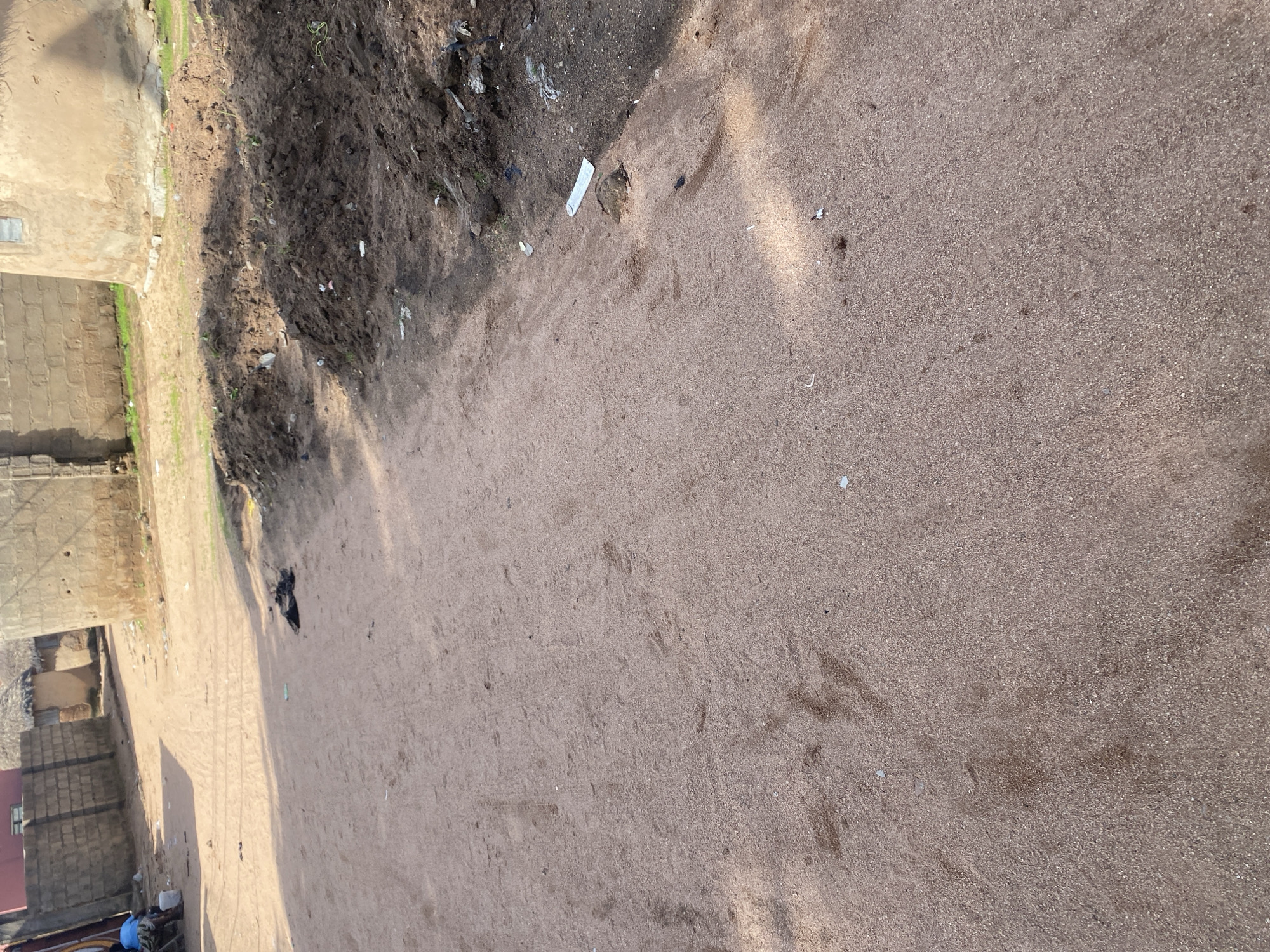 The image shows a sandy path in a dusty, rural or semi-rural environment. Buildings with thatched roofs and brick walls are visible in the background, suggesting a location in Nigeria. A person wearing a blue shirt and sitting on a bench is present on the left side of the path. A white bucket is near the person. Piles of dark soil are on the right side of the path, possibly indicating construction or gardening activities. Litter, including pieces of plastic and paper, is scattered on the ground and near the soil piles. The path shows tire tracks and footprints, indicating recent activity. The lighting suggests it is daytime, with shadows cast on the path.