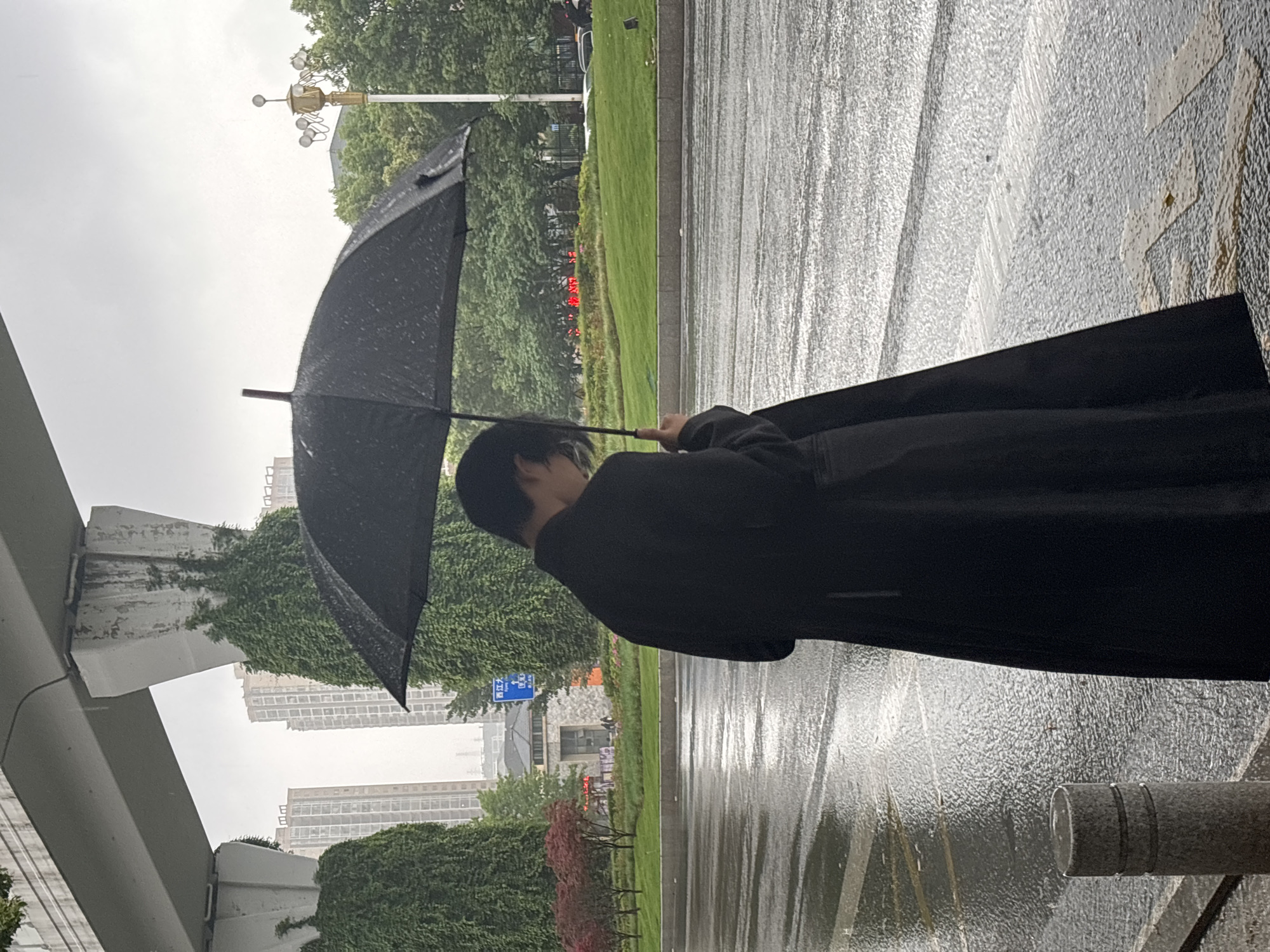 The photograph depicts an individual holding a black umbrella while standing on a wet pavement during a rainy day in Jiangjin, China. The person is wearing a long, dark coat. To the right of the subject is a wet street and a sidewalk. In the background, there is a line of green grass and trees, along with buildings and lamp posts. Raindrops are visible on the umbrella, indicating the intensity of the rainfall. A sign in Chinese characters is visible in the background. The overall impression is that of a typical rainy day in an urban environment.