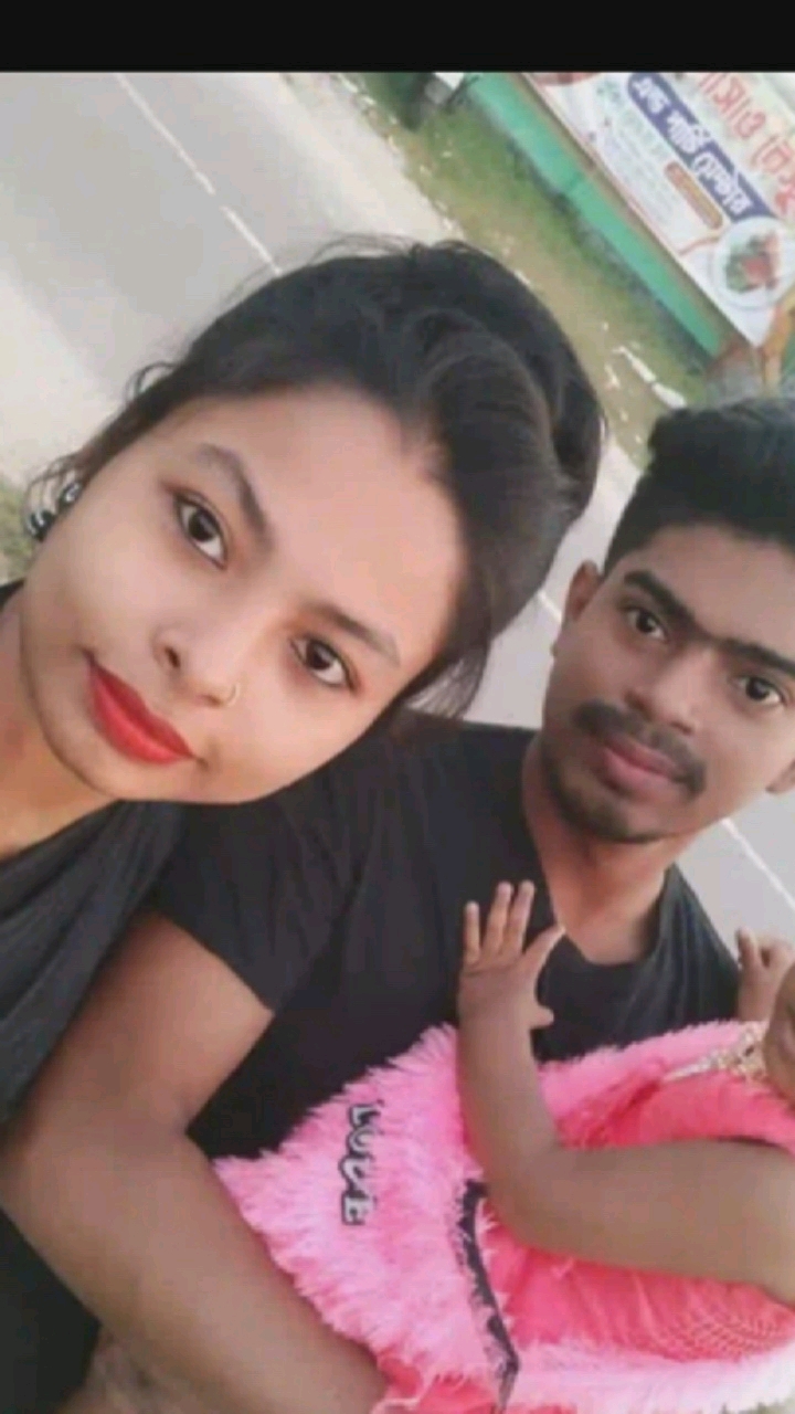 A portrait-style selfie captures three individuals, two adults and an infant, with a blurry outdoor background. The adult female on the left is facing the camera, with her head tilted. She has dark hair and is wearing dark clothing. To her right, the adult male is also looking towards the camera. He has dark hair and a mustache and is wearing a black t-shirt. The infant is held by the male, facing away from the camera and wearing a pink, fluffy outfit. The background appears to be a street scene with a sign on the right, though details are indistinct due to motion blur. The overall impression is a casual, informal photograph. The location is not specified as Hongkou District, China.