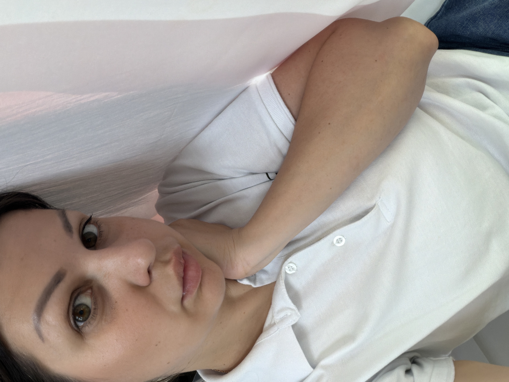 This is a close-up selfie of a woman taken indoors. She has dark hair and brown eyes, and is wearing a white polo shirt. Her left arm is raised, with her hand resting on her chin. The background appears to be a light-colored curtain. There are no discernible elements in the image that would indicate the location is Sochi, Russia.