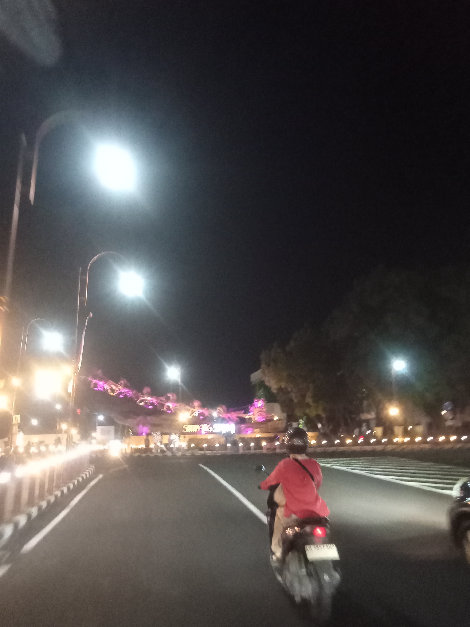 The image shows a nighttime scene on a road in Boyolali, Indonesia. A person is riding a scooter, viewed from behind. The rider is wearing a red shirt and a helmet. The road is paved and has white lane markings. On the left side of the road, there is a concrete barrier with black and white stripes. In the background, there are streetlights, buildings with decorative purple lighting, and trees. The sky is dark. The overall impression is a well-lit urban street at night.