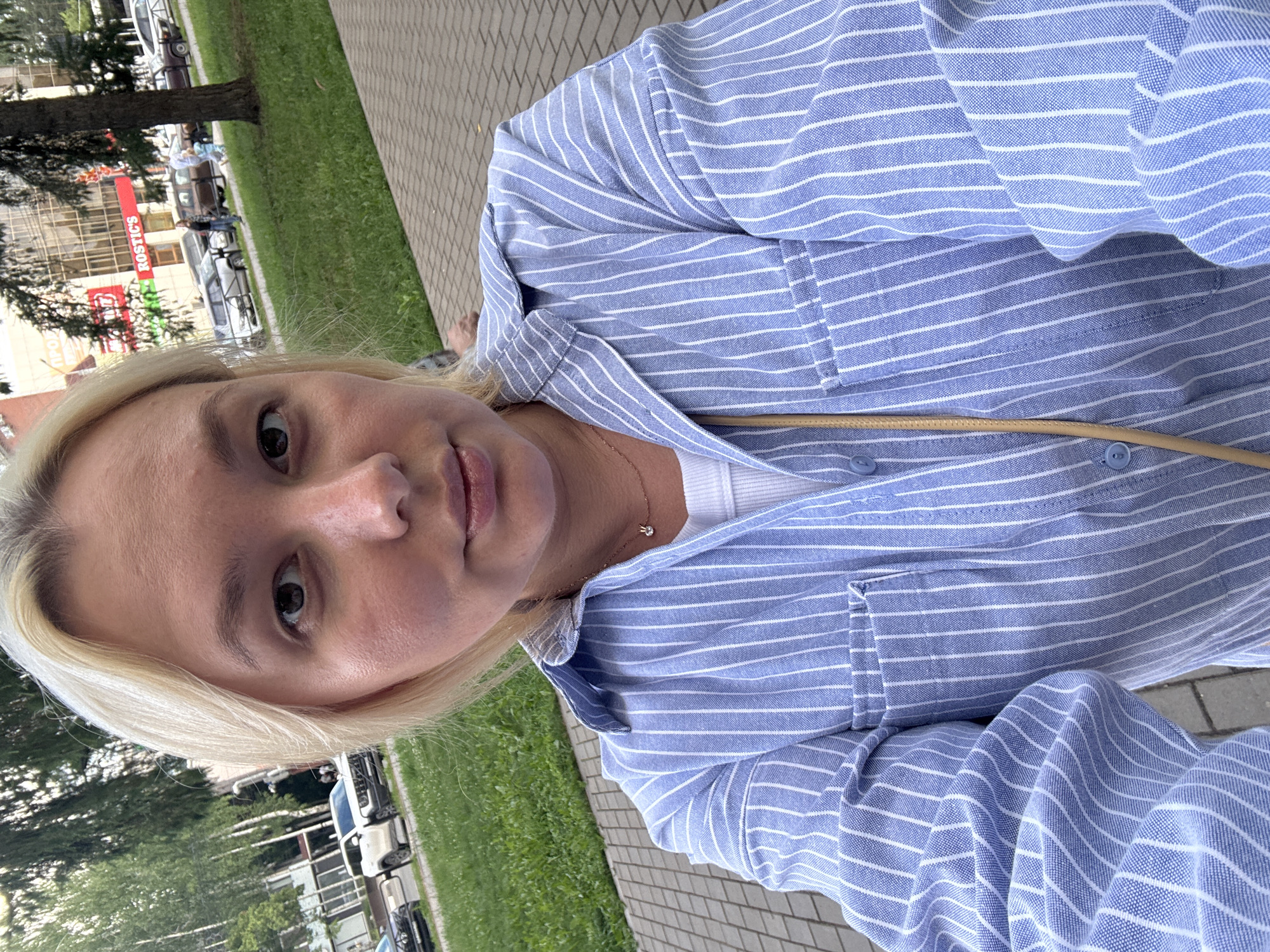 In Chaikovsky, Russia, a young woman with blonde hair takes a selfie outdoors. She is wearing a blue and white striped button-up shirt over a white tank top. A delicate gold necklace with a small pendant is visible around her neck. The background features a paved walkway, green grass, trees, and a building with the "ROSTIC'S" sign. Several cars are parked along the street, and a few people are walking in the distance. The overall scene suggests a casual daytime setting in an urban environment.
