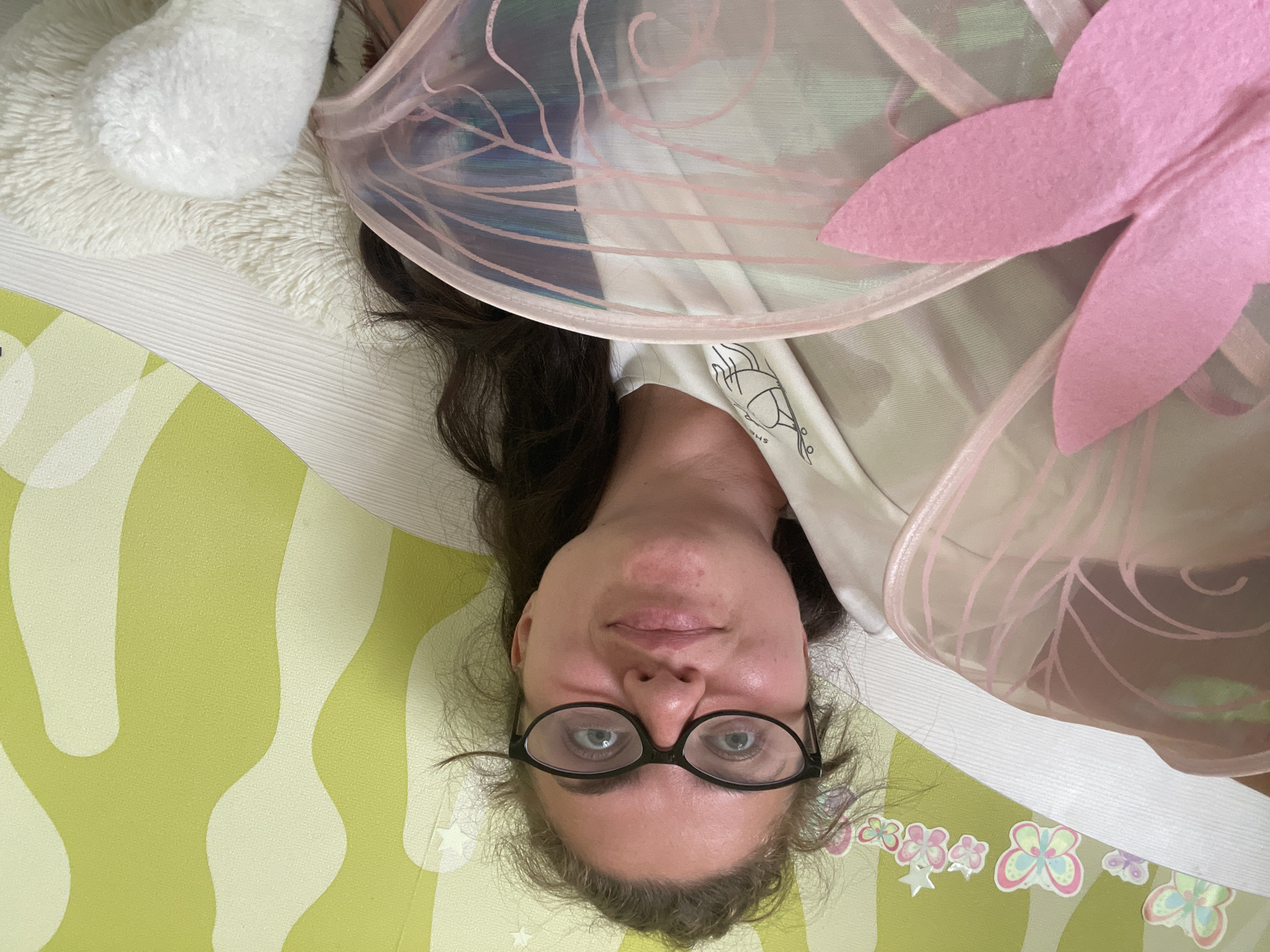A woman with dark hair, wearing glasses and a white t-shirt, lies on her back, possibly on a child's play mat featuring flower and butterfly designs. She is positioned under a translucent, pink-trimmed object resembling fairy wings with pink felt details. A white fluffy object rests to her left. The setting and location in the city of Moscow, Russia are not discernible from the given image.