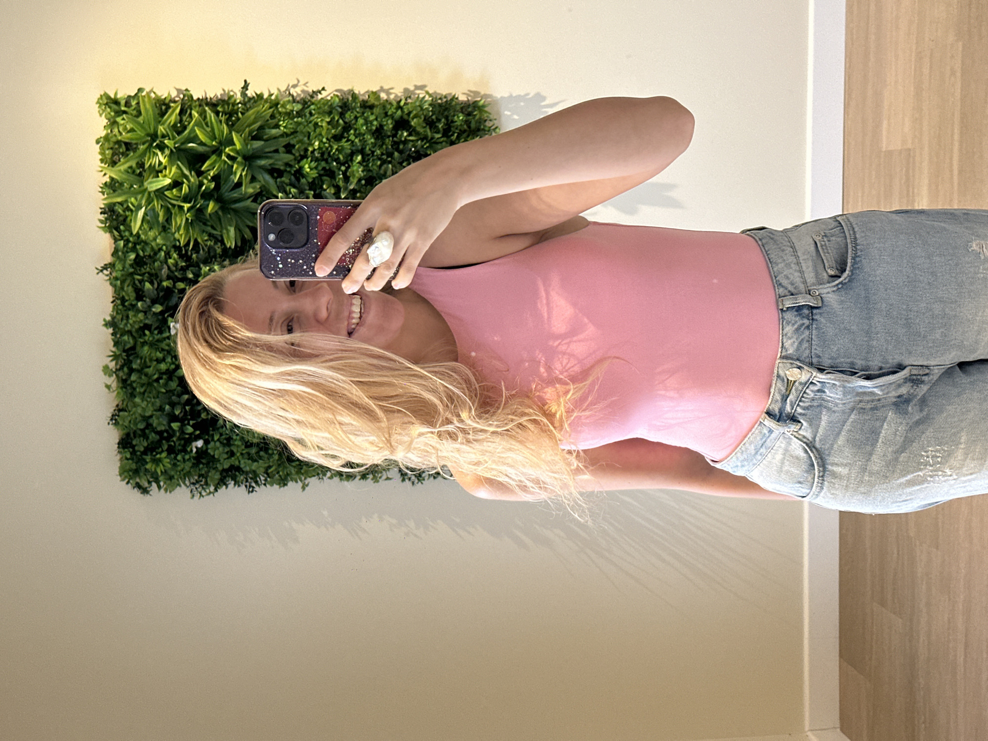 A young woman with blonde, wavy hair is taking a selfie in a room. She is smiling and looking at the camera, holding a smartphone with a starry, purple case. She is wearing a pink sleeveless top and light-wash, ripped denim jeans. On her right hand, she wears a large, irregular-shaped white ring. Behind her, a wall is decorated with a large, square panel of artificial green foliage. The wall to her left is a plain, light-colored surface with faint shadow patterns cast upon it, possibly from blinds or foliage. The floor is made of light brown wooden planks. The scene appears to be indoors, possibly in a fitting room or a home. There is no indication of the city of Reims, France, in this image.