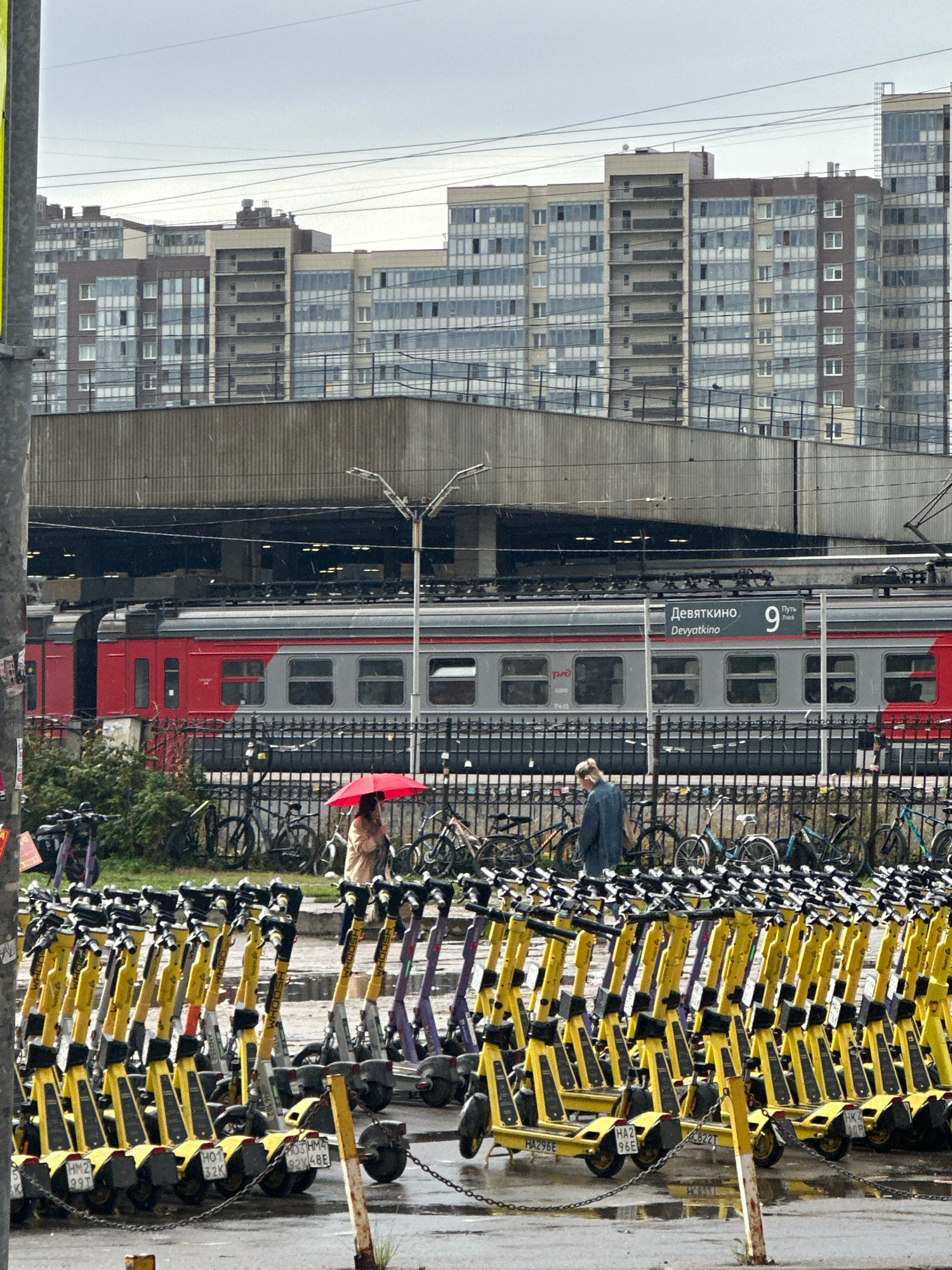 The image depicts a street scene in Novoe Devyatkino, Russia, characterized by a large train station with a red and gray train stopped at Track 9, as indicated by signage. In the foreground, a row of yellow electric scooters is parked, with some purple and other colored scooters also visible. Behind the scooters and a black metal fence, a person holding a red umbrella is walking, and another person in a blue jacket is standing. The background features a dense urban landscape of multi-story residential buildings under an overcast sky. The ground appears wet, suggesting recent rainfall.