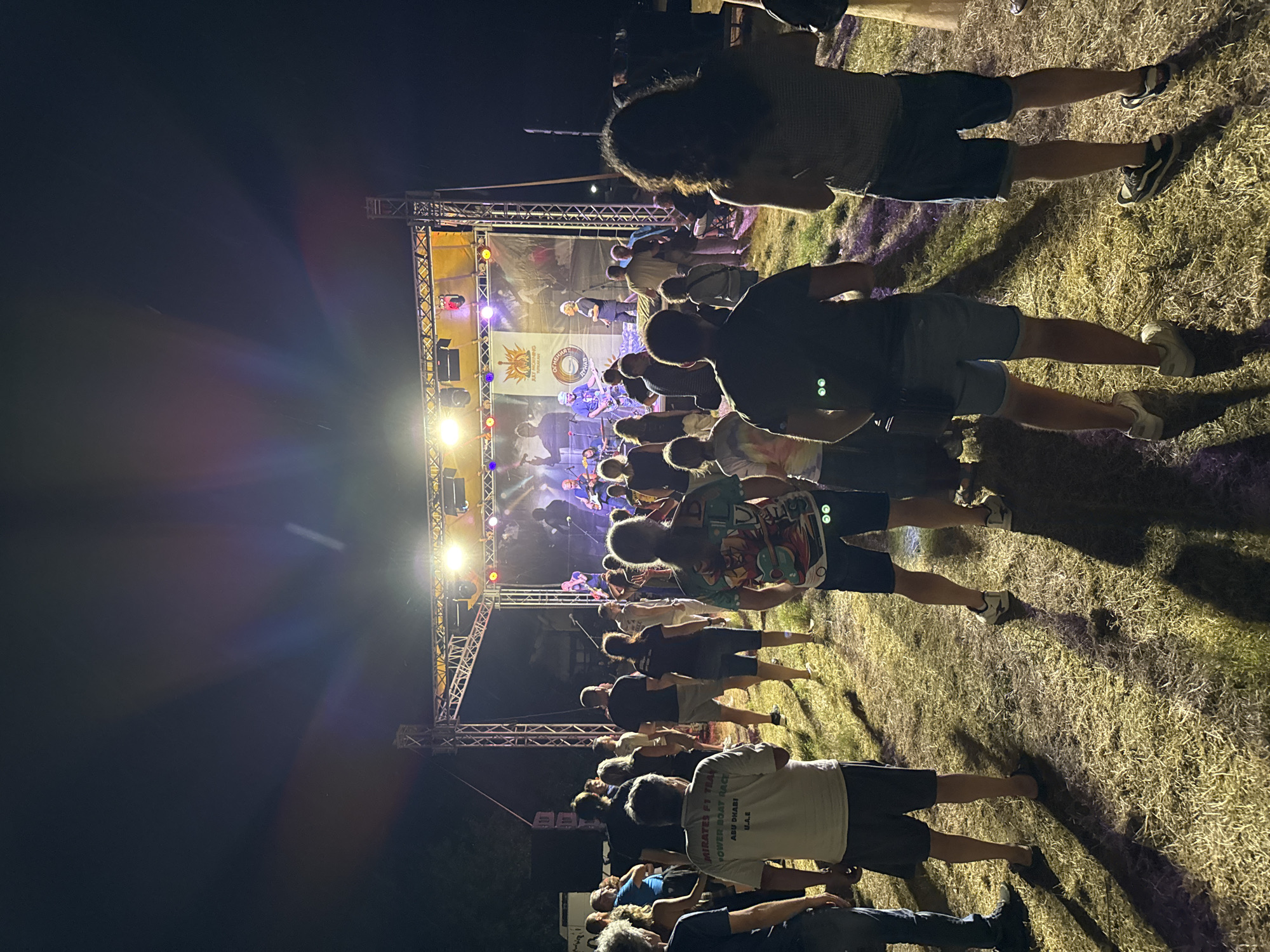 The image captures an outdoor concert event at night, set on a grassy field. A raised stage, illuminated by bright lights, is the focal point, featuring a band performing. Several people are visible in the foreground and midground, facing the stage and observing the performance. The audience appears to be a mix of adults, dressed casually in T-shirts, shorts, and jeans. The overall atmosphere suggests a lively gathering or festival. The event is located on the waterfront in the city of Tutrakan, Bulgaria.