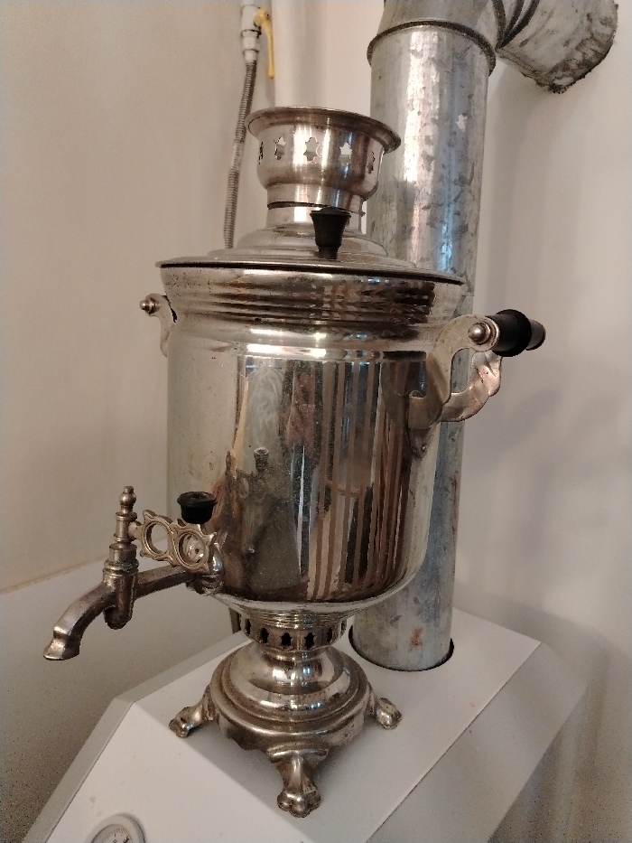An eye-level, indoor photograph captures a metallic samovar positioned on a white, angled surface in Lezhnevo, Russia. The samovar, appearing to be stainless steel or chrome-plated, exhibits a reflective, somewhat smudged surface. It features a decorative base with four ornate legs, a spigot with a black handle on its left side, and two handles with black grips on its upper body. A lid with a central black knob covers the main vessel, topped by a smaller, perforated crown.
In the background, a corrugated metal flue pipe rises vertically behind the samovar, then bends sharply to the right. To the upper left, a white vertical pipe is affixed to a light-colored wall, from which a flexible metal hose extends, terminating in a yellow-handled gas shut-off valve. The white surface beneath the samovar appears to be a utility platform or the top of a mechanical unit, with a partial view of a circular gauge visible at the bottom left edge of this platform. The overall lighting suggests an interior setting.