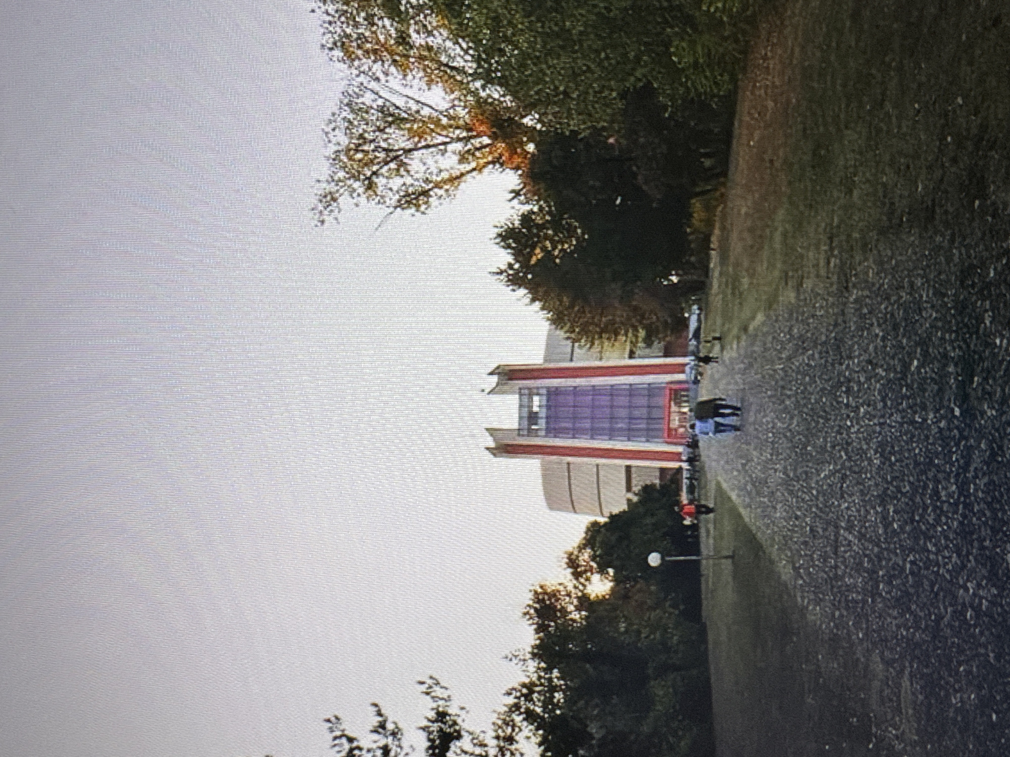 The image captures an exterior view of a modern building with a prominent glass facade, possibly a cultural or educational institution, situated in Varna, Bulgaria. The building features a central section with vertical red accents flanking a large purple-tinted glass structure. The approach to the building is a wide, gravel-covered pathway lined with grass and trees. Several individuals are visible walking on the pathway. Two people are walking away from the camera towards the building, appearing to be in conversation. Further down the path, another individual is walking alone. The scene is set against a backdrop of trees and a clear, light-colored sky, suggesting it is daytime. The overall impression is one of a public space with people enjoying the outdoors.