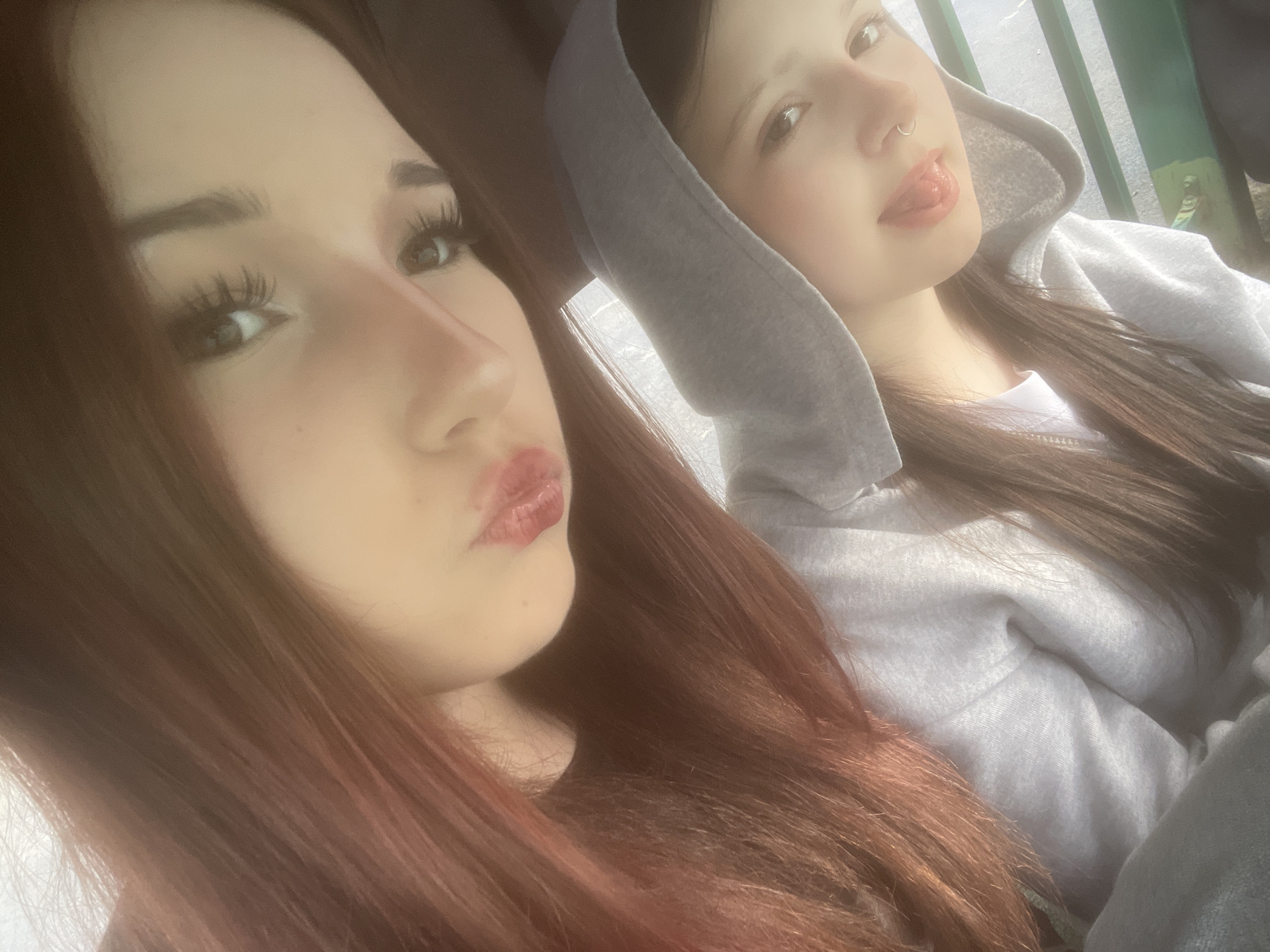 Two young women are captured in a close-up, casual photograph, likely a selfie. The woman in the foreground, positioned to the left, has long, reddish-brown hair that falls over her shoulder. She has prominent dark eyebrows and long, dark eyelashes. Her lips are a glossy red, puckered into a "kissy face" gesture.
Behind her, partially visible to the right, is another young woman. She is wearing a grey hooded sweatshirt with the hood pulled up. Her dark hair is visible beneath the hood. She has a nose piercing, a silver ring in her septum, and is playfully sticking her tongue out. Her expression is lighthearted and relaxed.
The background is softly blurred but shows hints of an outdoor or semi-outdoor setting, with vertical green bars or fencing visible behind the second woman, suggesting railings or a barrier. The lighting appears bright and natural, indicating it is daytime. The overall mood of the image is informal and friendly, capturing a candid, playful moment between the two individuals. There is no visible text in the image. The location context for this scene is Saint Petersburg, Russia.