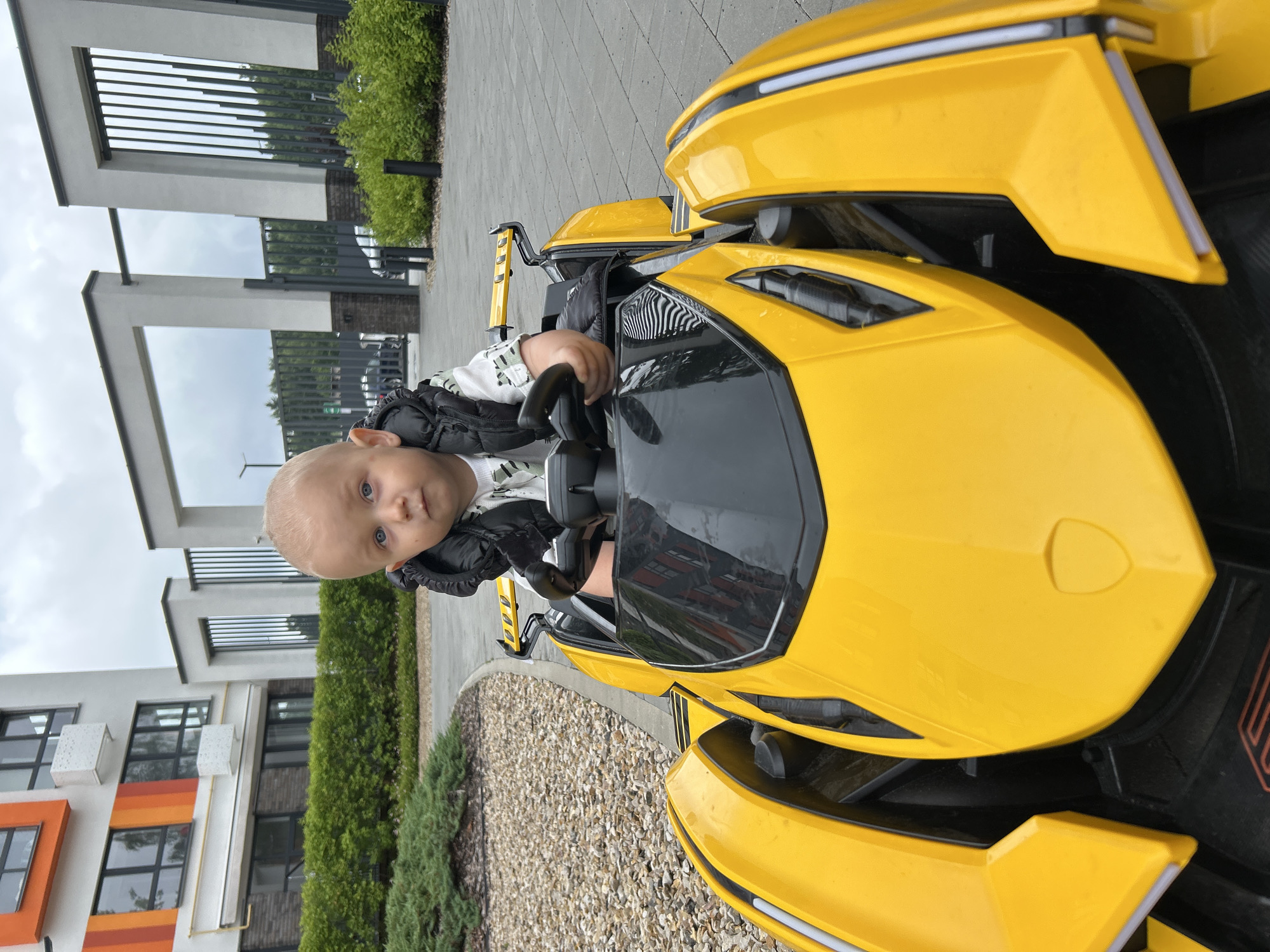 A young child with blonde hair and blue eyes is seated in the driver's seat of a yellow toy car. The child is wearing a black vest over a patterned shirt and is holding the steering wheel. The toy car has a black windshield and accents. In the background, there is a modern building with orange and white geometric patterns, lush green bushes, and a paved walkway. The scene appears to be in an outdoor urban setting, possibly a courtyard or residential area. The sky is overcast.