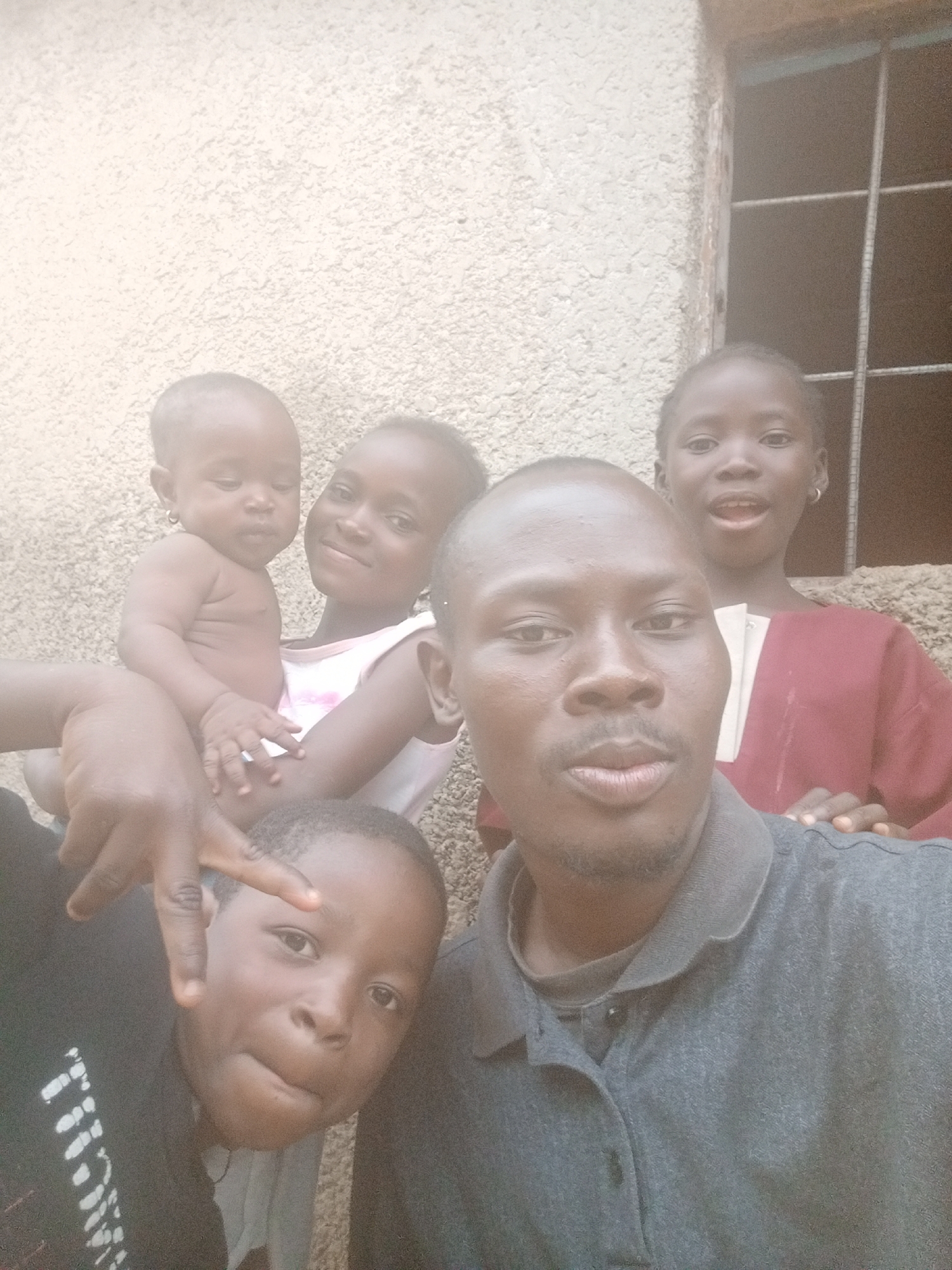 A selfie depicts a man and four children in Sabon Tasha, Nigeria. The man, positioned in the foreground, has dark skin, a shaved head, and a thin mustache. He wears a gray polo shirt. A young boy in a black shirt is to his left, making a hand gesture. An infant with earrings is held by a girl in a light pink tank top. Another girl in a reddish-brown top stands behind the man, near a window with bars. The background is a beige, textured wall.
