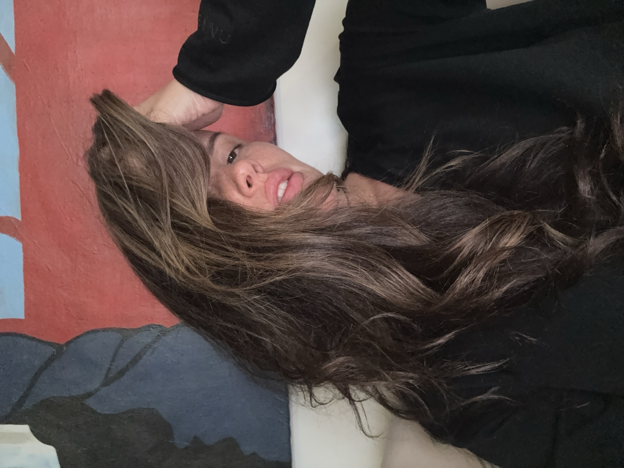 A woman is lying down, angled slightly towards the camera. Her head rests on her hand, displaying brunette hair with lighter highlights. She wears a black top. Behind her is a section of a painting or wall with a blue square on a red background and a darker blue section below. The scene does not provide sufficient information to determine the location in Krasnoyarsk, Russia.