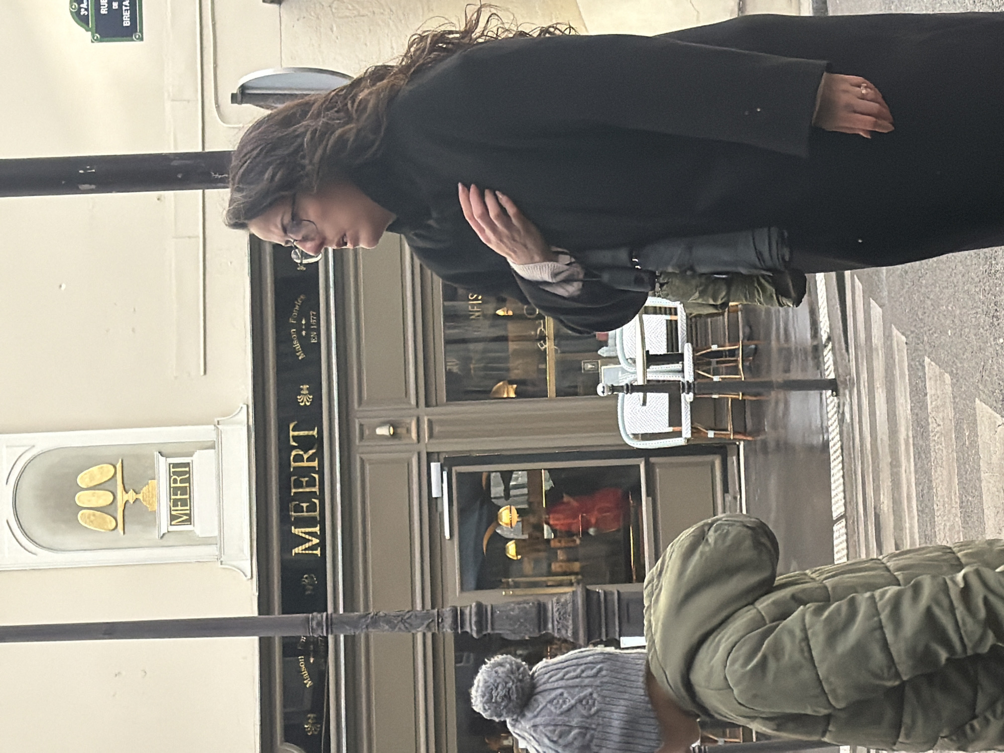 The scene depicts a street in Paris, France, specifically outside the "MEERT" patisserie, identifiable by its prominent golden signage and a partial street sign indicating "RUE DE BRETA," signifying Rue de Bretagne. The street is wet, suggesting recent precipitation.
In the foreground, a woman with dark, wavy hair and glasses stands facing right, looking upwards with her mouth slightly open. She is wearing a long, dark (likely black) coat, with her left hand resting on her chest, revealing a light-colored sleeve. A folded dark umbrella is held under her left arm. A ring is visible on her right ring finger. To her left, partially in the frame, is a person (likely a child) seen from the back, wearing a grey knitted beanie with a pompom and a green quilted puffer jacket. This individual is positioned on a pedestrian crosswalk, which extends across the wet asphalt.
A dark, ornate lamppost stands between the two individuals. The "MEERT" patisserie facade in the background is light-colored, featuring a central arched niche with three golden oval shapes on a pedestal above the "MEERT" name. The storefront itself is dark brown with large glass windows, reflecting warm interior lights and a red object. The shop's full name "MEERT" is displayed in golden letters above the entrance, with "Maison Fondée EN 1877" also visible. To the right of the shop, two white outdoor chairs and small tables are present.