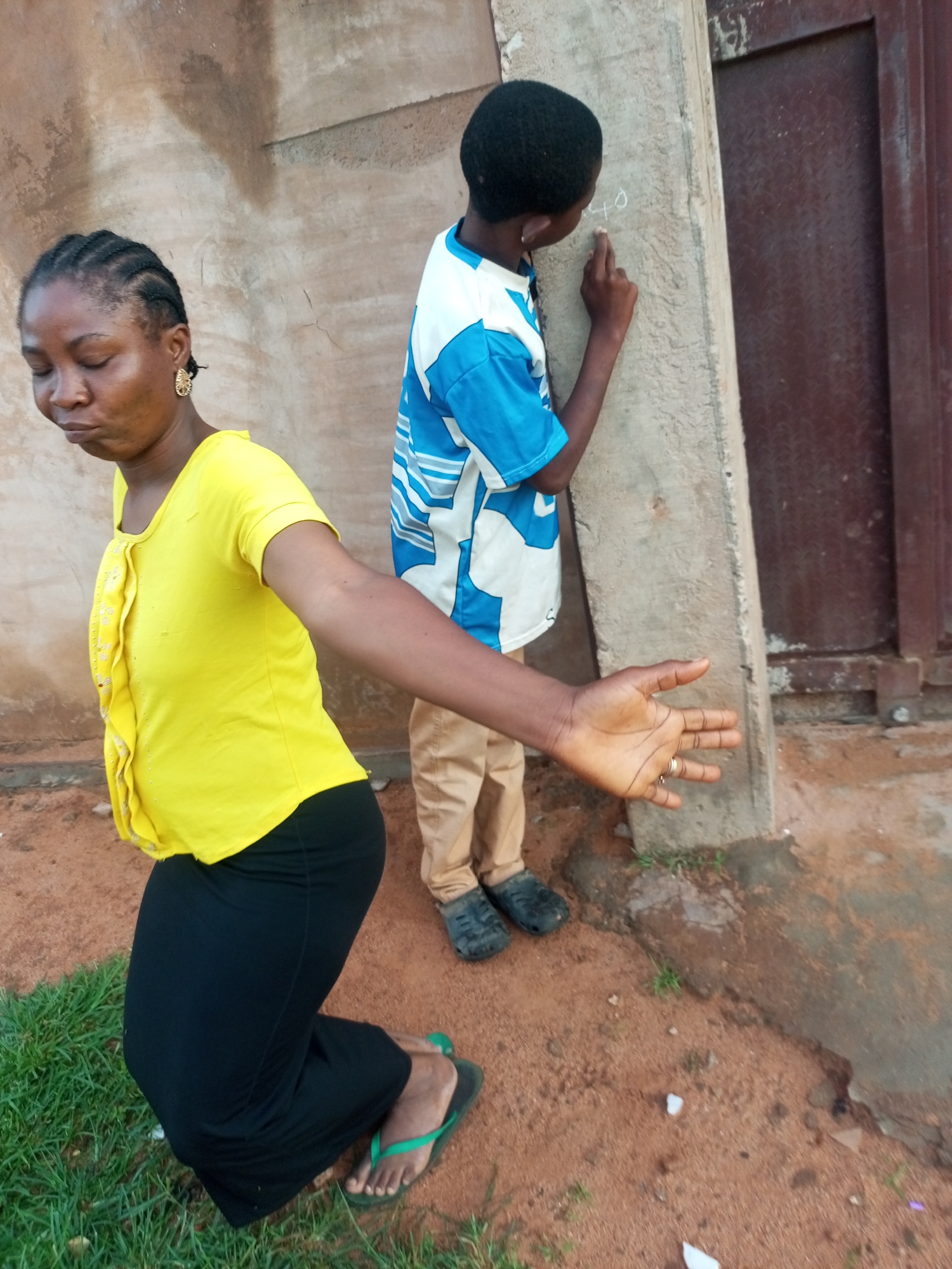 In Tempure, Nigeria, a woman in a bright yellow top and black skirt kneels on the ground with her hand outstretched. Her eyes are closed and her head is bowed, suggesting a moment of prayer or contemplation. Beside her, a young boy dressed in a blue and white patterned shirt and khaki pants stands by a concrete pillar, his back to the viewer. He is engaged in drawing with chalk on the pillar, with the number "40" visible. The ground is a mix of reddish-brown soil and patches of green grass. In the background, a dark brown, corrugated metal door stands against a textured, light brown wall. The scene appears to be outdoors, possibly in a residential or communal area.