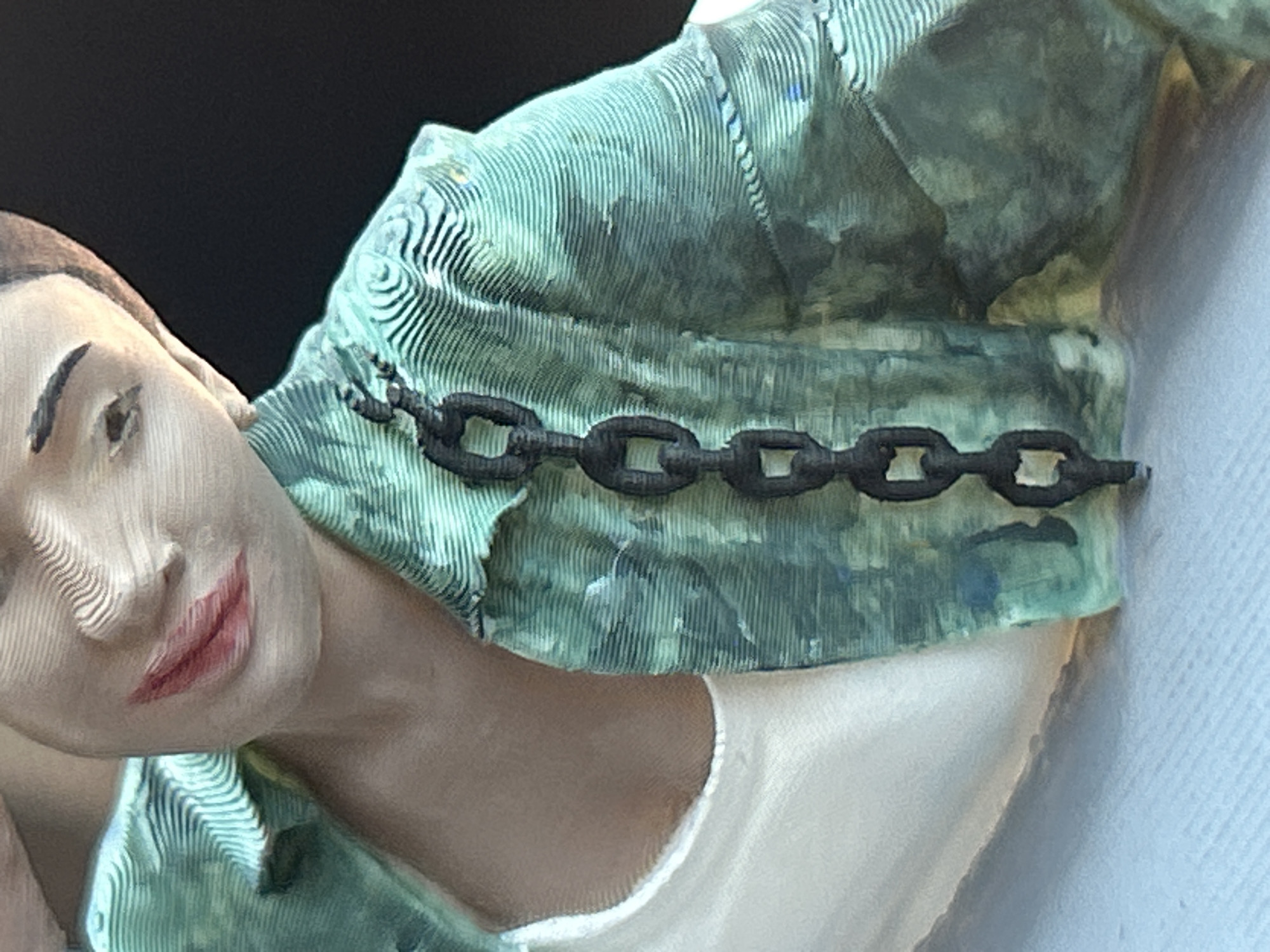 The image is a close-up of a 3D printed figurine of a person. The figurine is wearing a green patterned shirt over a white undershirt, and a black chain hangs from the shirt. The figurine has red lipstick and is partially obscured by the dark background. There is no discernible location in the city of Lozen, Bulgaria.