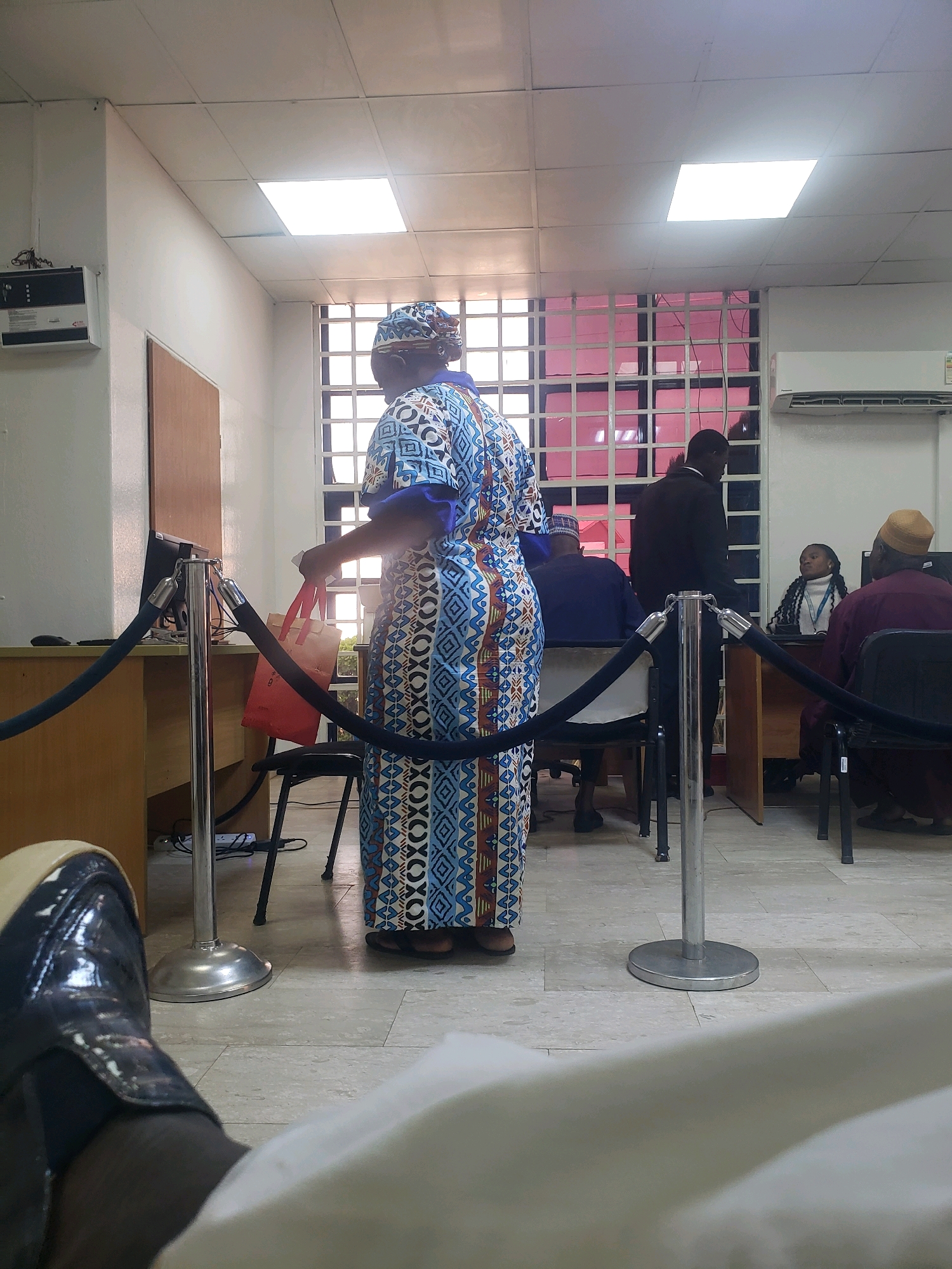 The image depicts an indoor scene, likely a government office or a bank in Kakuri Gwari, Nigeria. In the foreground, the legs and shoes of a person are visible, suggesting they are sitting or standing out of frame. In the mid-ground, a woman wearing a colorful patterned dress and headwrap stands facing away from the camera, holding a red bag. She is positioned near a counter with a computer monitor. Behind her, a barred window with pink panes offers a view of the outside. Several other individuals are present, including a man in a suit standing and facing the counter, and two individuals, one wearing a tan cap and the other a maroon garment and tan cap, seated at desks. A young woman in a white turtleneck is also seated at a desk. The environment is functional, with fluorescent lighting, air conditioning units, and stanchions with ropes indicating a queue. The overall impression is of a busy service area.