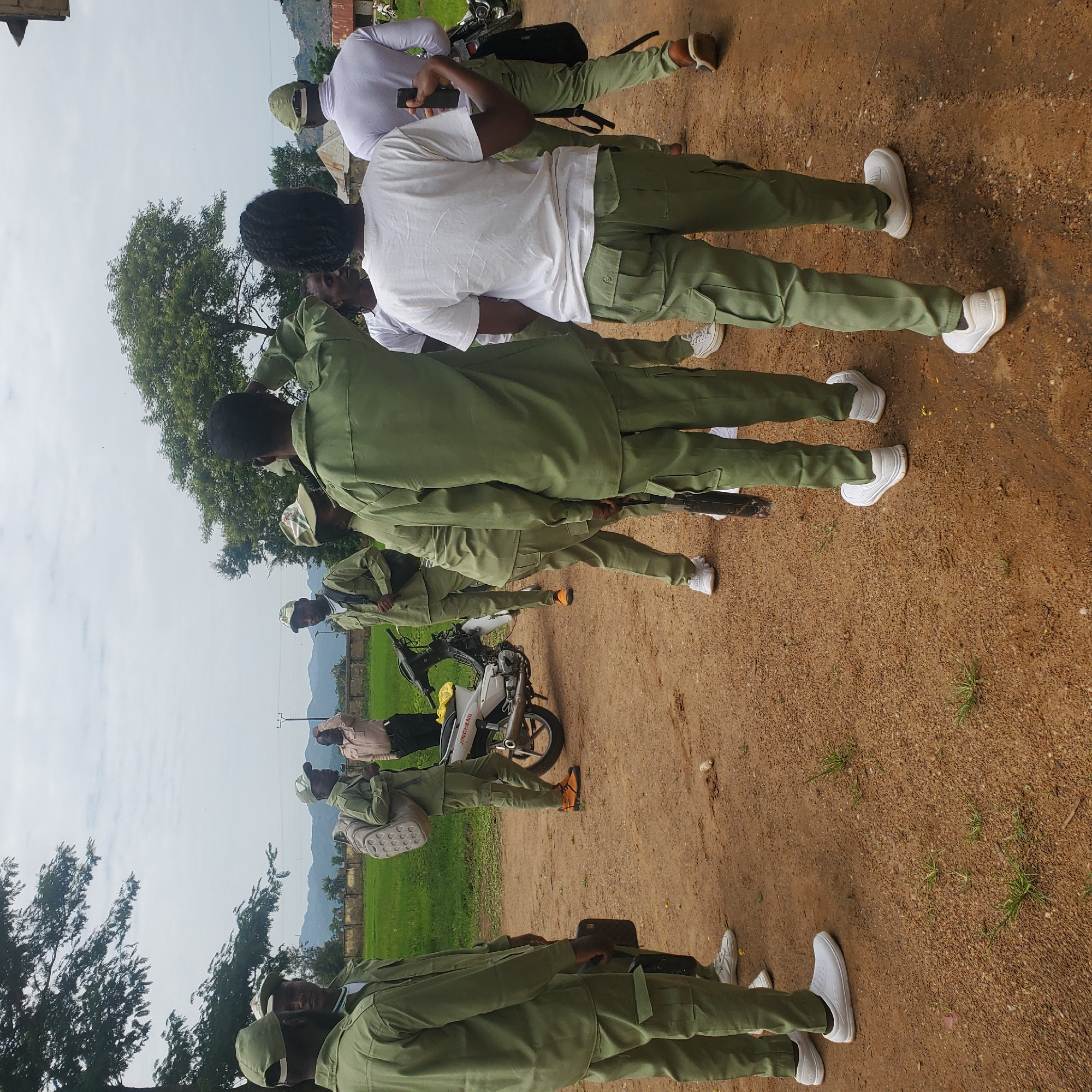 In Toro, Nigeria, a group of individuals, predominantly wearing light green uniforms and caps, are gathered outdoors on a dirt path. The group appears to be in an informal huddle, with several people facing away from the camera and others engaged in conversation. A motorcycle is parked on the right side of the frame. In the background, there is a grassy field, a low stone wall, and trees under a cloudy sky. The overall setting suggests a rural or semi-rural environment. The people are dressed in matching outfits, indicating a possible shared affiliation, such as a youth corps or a uniformed organization. The individuals are of various ages and genders, and some are carrying bags or backpacks.