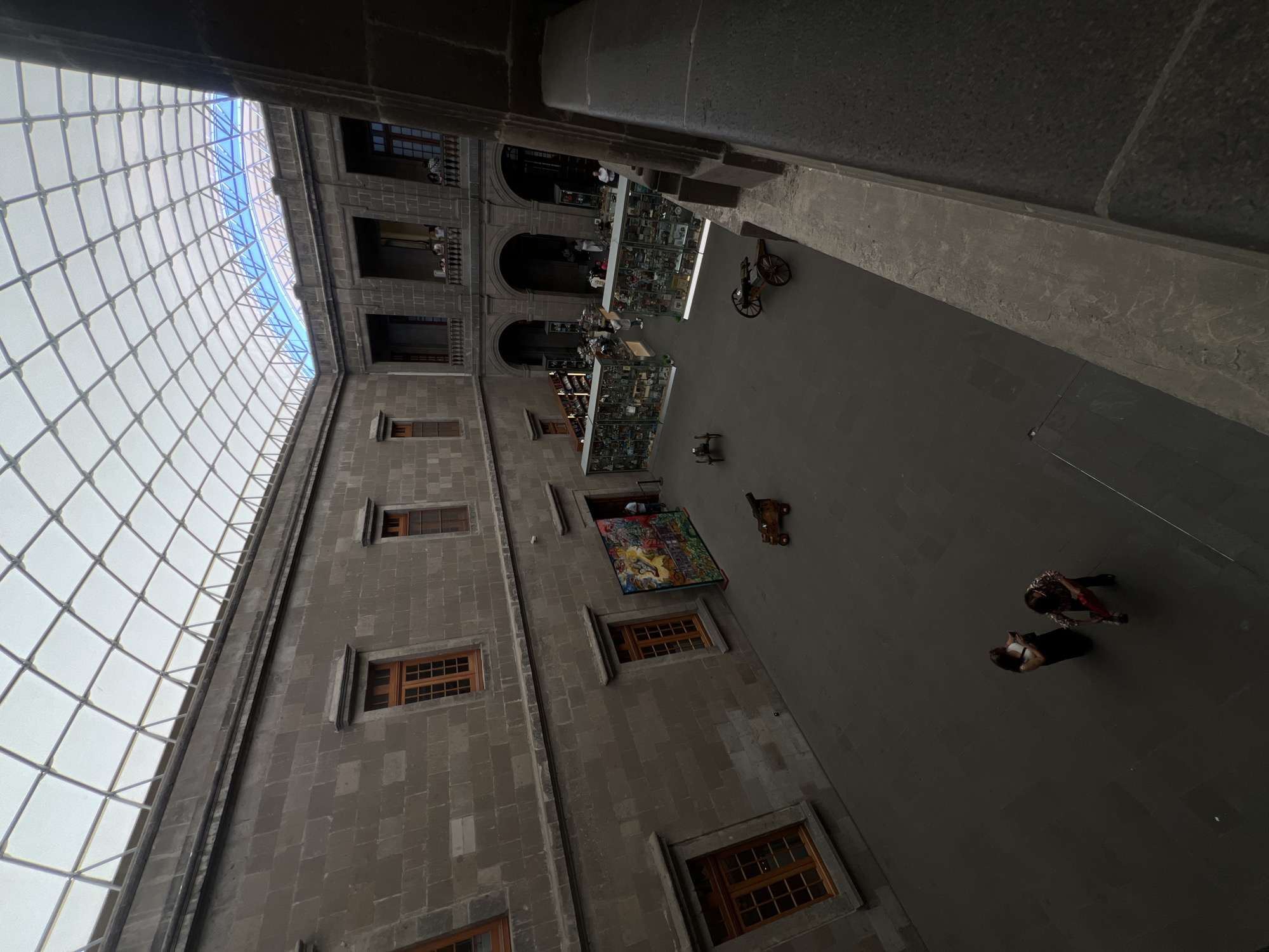 The image depicts an interior courtyard in Mexico City, Mexico. The courtyard is enclosed by a building with multiple stories, featuring windows and balconies. A translucent grid-like structure covers the courtyard, allowing diffused light to enter. In the courtyard itself, display cases contain various objects, possibly artifacts or exhibits. Several individuals are visible, some near the display cases and others walking in the open area. The overall environment suggests a museum or historical site.