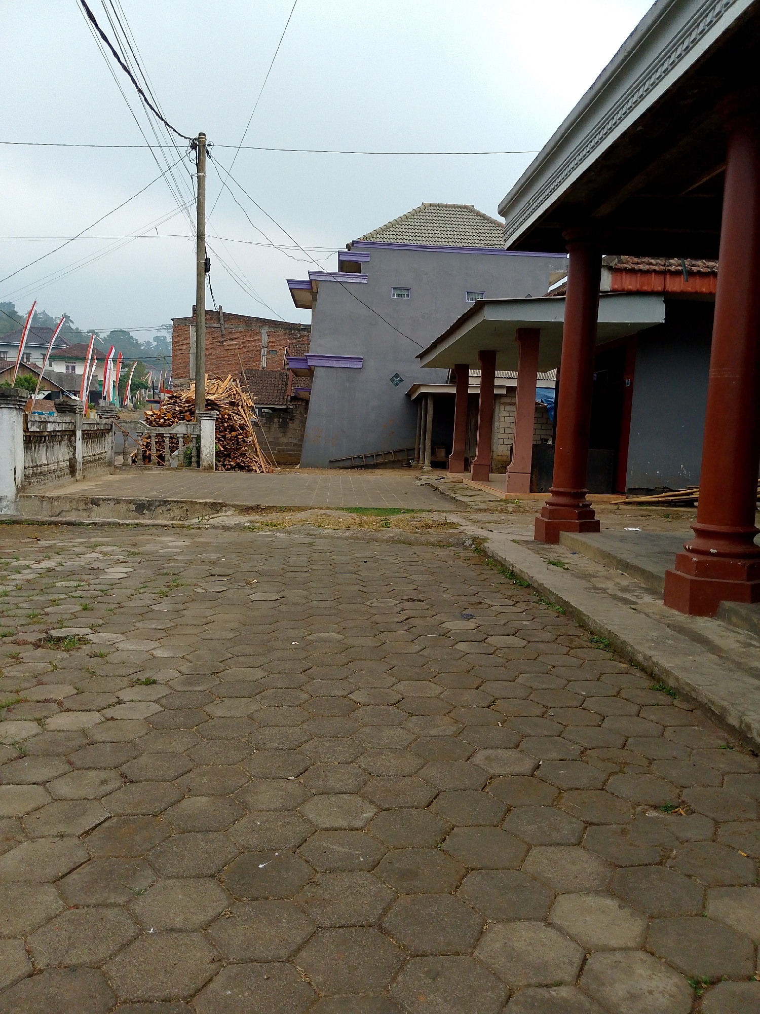 The image depicts an outdoor scene in Babadan, Indonesia. In the foreground, a paved area is covered with hexagonal concrete interlocking pavers, some of which are overgrown with sparse grass. A raised concrete curb runs along the right side of this paved area, with a gutter between it and a building with large, reddish-brown cylindrical pillars supporting a covered walkway. The building's exterior walls are a dark gray color, and the roofline features decorative trim.

Beyond the walkway, a pile of cut wood is stacked next to a brick building. A concrete wall with a decorative balustrade separates this area from a street or pathway where several Indonesian flags, red and white, are displayed on poles. Further in the background, more residential buildings and lush green hills are visible under a cloudy sky. A utility pole with a dense network of electrical wires stands prominently in the middle ground. The overall impression is of a residential or communal area with visible infrastructure and preparations, possibly for an event given the flags.