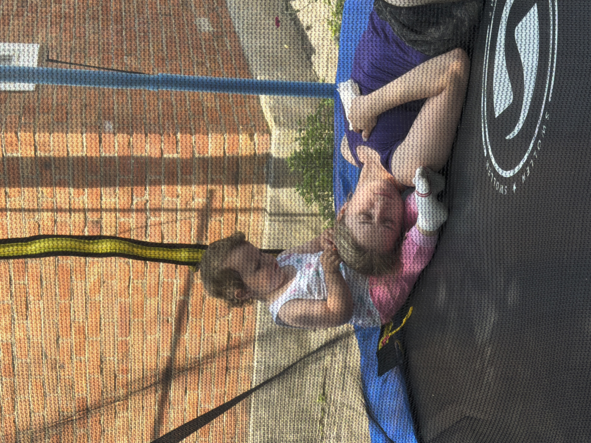 In the city of Komsomolskiy, Russia, two individuals, a younger child and an adult, are lying on a black trampoline. The trampoline has a safety net enclosure made of woven material, with visible poles. The background consists of a brick wall with sunlight casting shadows, and some greenery and a flower. The adult, lying on their back with their head resting on their arm, wears a purple top and dark shorts. The child, sitting up and leaning on the adult, is wearing a white floral-patterned top and pink bottoms. The child appears to be interacting with something in their hands. The overall scene suggests a casual, outdoor activity.