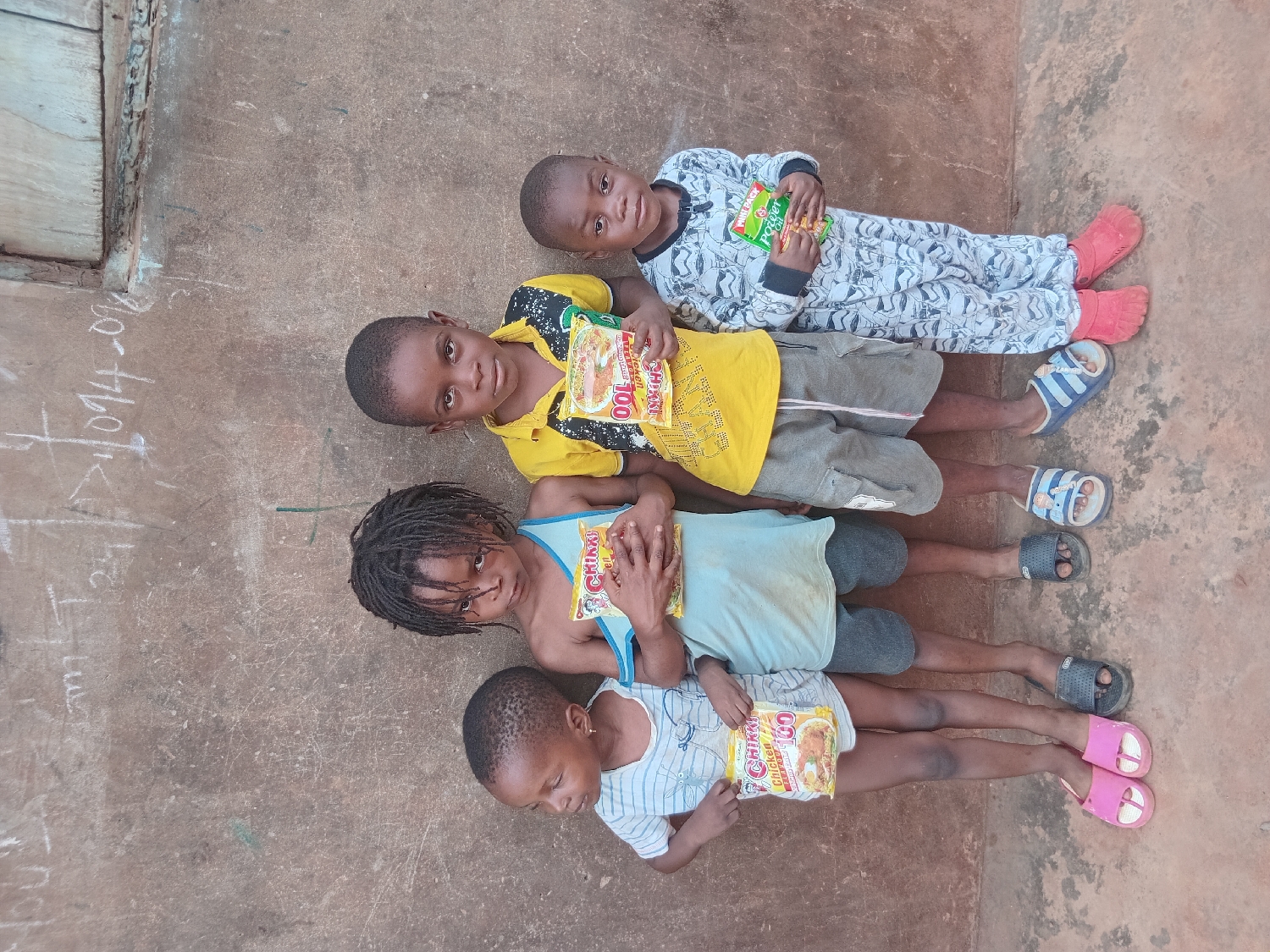 Four young children are depicted lying horizontally on a reddish-brown concrete surface, with their heads positioned towards the left side of the frame and their feet towards the right. The setting appears to be an outdoor paved area, brightly lit, suggesting daytime.
From left to right in the image:
The leftmost child, with dreadlocks, wears a light blue tank top and dark shorts. They hold a white and yellow packet labeled "CHING'S" and wear dark slip-on sandals. They are looking directly at the camera.
Next to them, a child in a striped short-sleeved shirt and dark shorts holds a white and yellow packet, "CHING'S CHICKEN 100," and wears pink slip-on sandals. This child is looking downwards.
Further to the right, a child wearing a yellow t-shirt with black patterns and grey shorts holds a yellow packet marked "CHING'S CHICKEN 100." They wear blue and white striped sandals and are looking directly at the camera.
The rightmost and youngest child wears a patterned, long-sleeved jumpsuit and red slip-on shoes, holding a green packet labeled "Power G." This child is also looking directly at the camera.
Each child is holding a packet of what appears to be instant noodles or a similar food item. In the upper left corner of the frame, an old, weathered wooden doorframe or door edge is visible. Markings resembling "T 24" and "094 - 016" are faintly visible, possibly scratched or chalked onto the concrete surface in the upper left background. The overall scene suggests a simple, everyday outdoor environment in Shagamu, Nigeria.