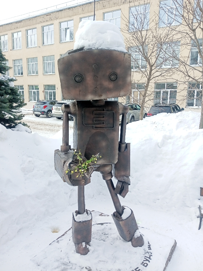 A static, full-body outdoor shot depicts a dark metallic-colored robot statue standing in a deeply snow-covered environment in Chaikovsky, Russia. The robot, approximately human height, features a rectangular head topped with a significant accumulation of white snow, two circular indentations serving as eyes, and a prominent rectangular recess on its chest containing an 'E' or 'ш' shaped emblem. An antenna-like rod extends from its head. Its left arm, angled forward, holds a small bouquet of green sprigs adorned with numerous tiny star-shaped white and yellow ornaments. The robot stands on a flat base, partially obscured by snow, where Cyrillic text is visible. In the background, a large, light-colored multi-story building with numerous rectangular windows, indicative of an institutional structure, stands under an overcast sky. A paved area partially cleared of snow, located between the robot and the building, shows several parked vehicles, including a grey crossover SUV on the left and a darker SUV further right. To the immediate left of the statue is a large, snow-laden coniferous tree, while several bare deciduous trees are visible in front of the building. The entire foreground and much of the mid-ground are covered in undisturbed white snow, characteristic of a winter setting.