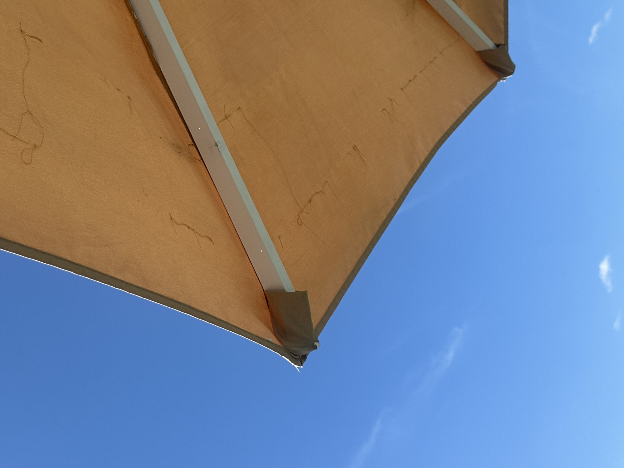 An outdoor patio umbrella made of tan fabric is angled against a clear, bright blue sky. The umbrella's frame, visible underneath the fabric, appears to be white or light gray metal. The fabric itself shows some marks or stains, possibly from water or dirt. A thin wisp of a cloud is present in the upper left quadrant of the sky, and a smaller, puffier cloud is visible near the bottom right. The scene is observed from below, looking up at the umbrella and the sky. There are no people or animals visible in the image. The setting is likely a sunny day, potentially in a park or outdoor seating area. The location is identified as Khotianivka, Ukraine.
