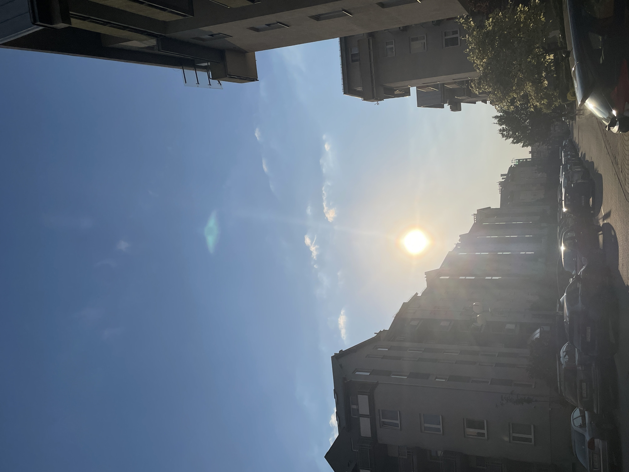 The image shows a street scene in an urban environment. The buildings are high and appear to be residential or commercial. Parked cars line the street on the right side of the frame. The sun is visible in the sky. The sky is mostly blue with a few thin clouds. The specific location of Unknown, Unknown, cannot be determined from the image. No people are visible.