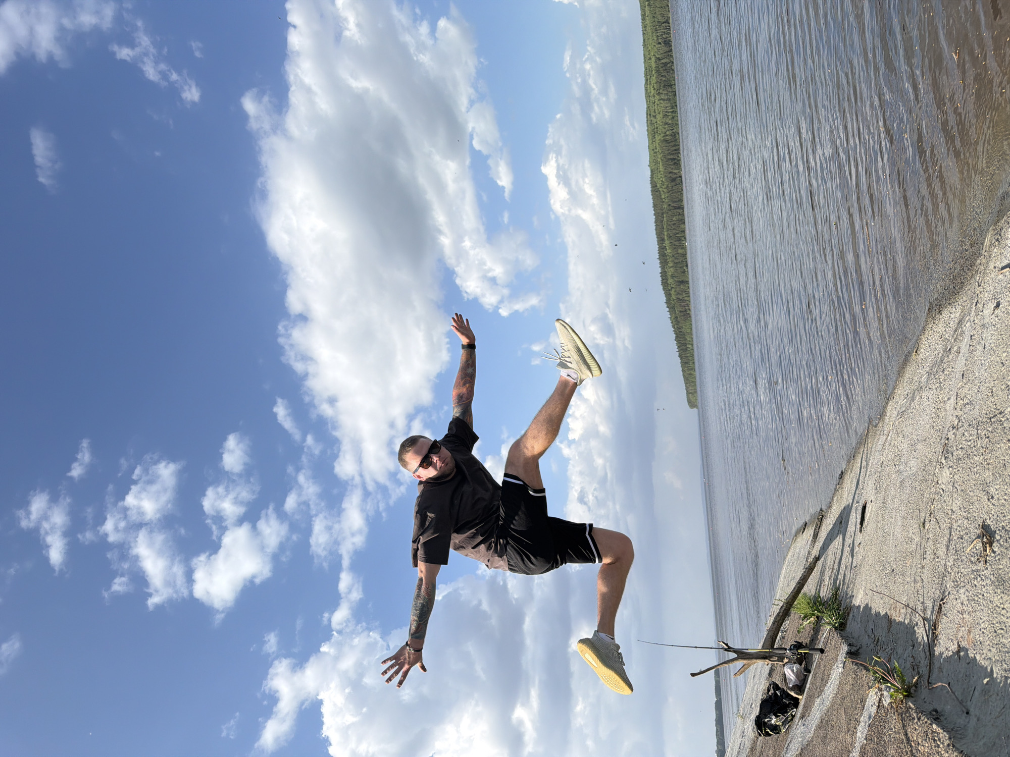A man is performing a split jump on a concrete structure, with a body of water and a tree line in the background, and a blue sky with clouds in the top left. The man is wearing a black t-shirt, black shorts with white stripes, sunglasses, and light-colored athletic shoes. He has tattoos on both arms. The concrete structure has grass and vegetation growing from it, along with a fishing pole and a bag. The image is taken at an angle, with the man appearing diagonal in the frame. Based on context clues (Chaikovsky, Russia), the location is likely near the Kama River or a smaller body of water in that area.