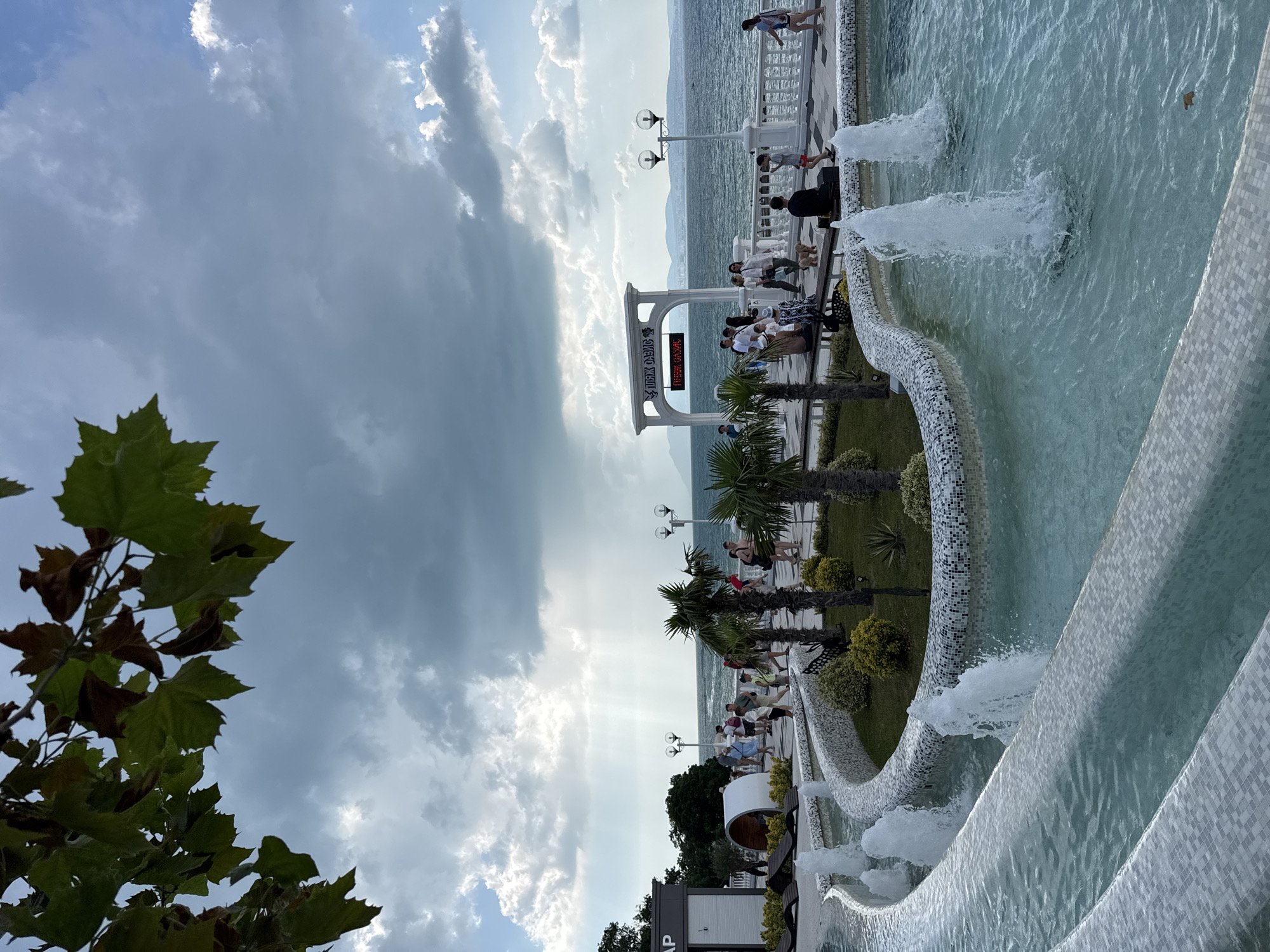 The image depicts a waterfront promenade in Novorossiysk, Russia, featuring a multi-tiered fountain in the foreground. The fountain's curves are lined with white and grey mosaic tiles, and several water jets are active, creating a lively aquatic display. Beyond the fountain, a paved walkway runs parallel to the sea, with a low decorative wall separating it from a landscaped area.
This landscaped area contains small palm trees, green bushes, and scattered plants. A cylindrical structure, possibly a modern seating area or pavilion, is visible on the left. People are seen strolling along the walkway, some in groups, others individually, engaging in typical promenade activities. One person is seated on a bench. A small dog is also present.
An arched white structure with signage in Cyrillic characters stands further down the promenade, framing the view of the sea and the distant, hazy coastline. Lampposts are placed along the walkway. The sea is a deep blue, and the sky above is a mix of dramatic grey clouds and patches of blue sky, with sunbeams piercing through the clouds in the distance, suggesting a dynamic weather pattern. Foliage from a tree hangs into the upper left corner of the frame.