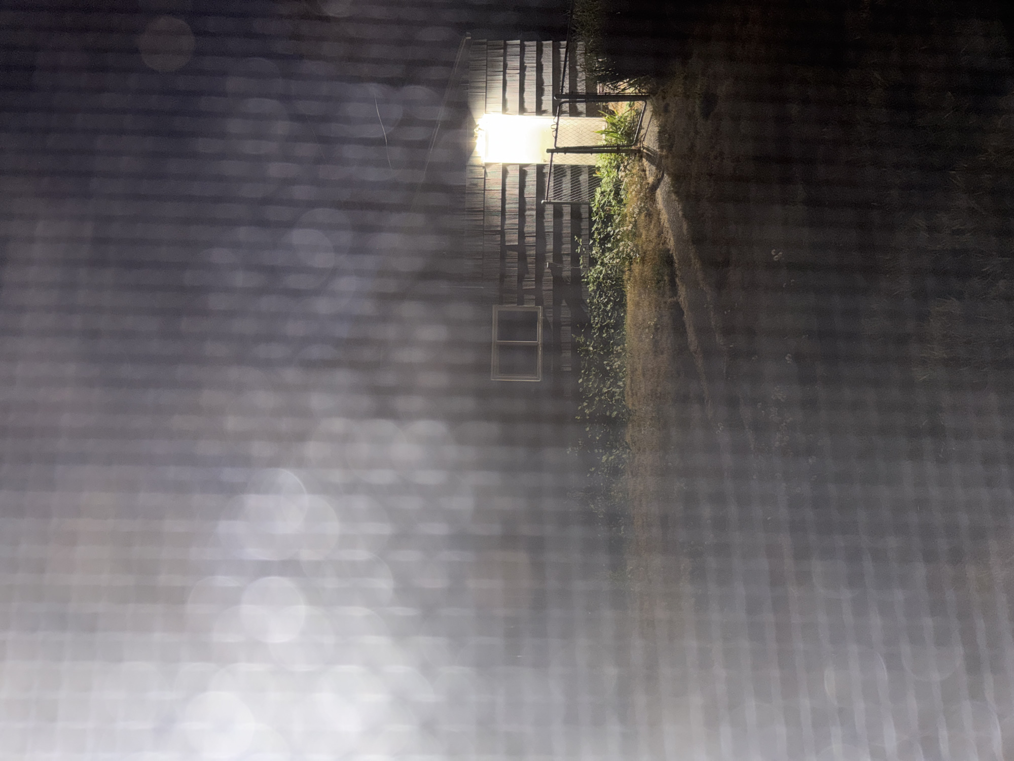 An outdoor, nighttime photograph depicts a small, wooden building with horizontal siding and a brightly lit doorway. A single window is visible on the left side of the building. The structure is surrounded by sparse vegetation and a chain-link fence with an open gate. A pathway or driveway leads towards the building. The foreground is obscured by a blurry, netted overlay, possibly a screen or mesh, with bokeh effects from a light source. The overall impression is that of a somewhat isolated and dimly lit scene.