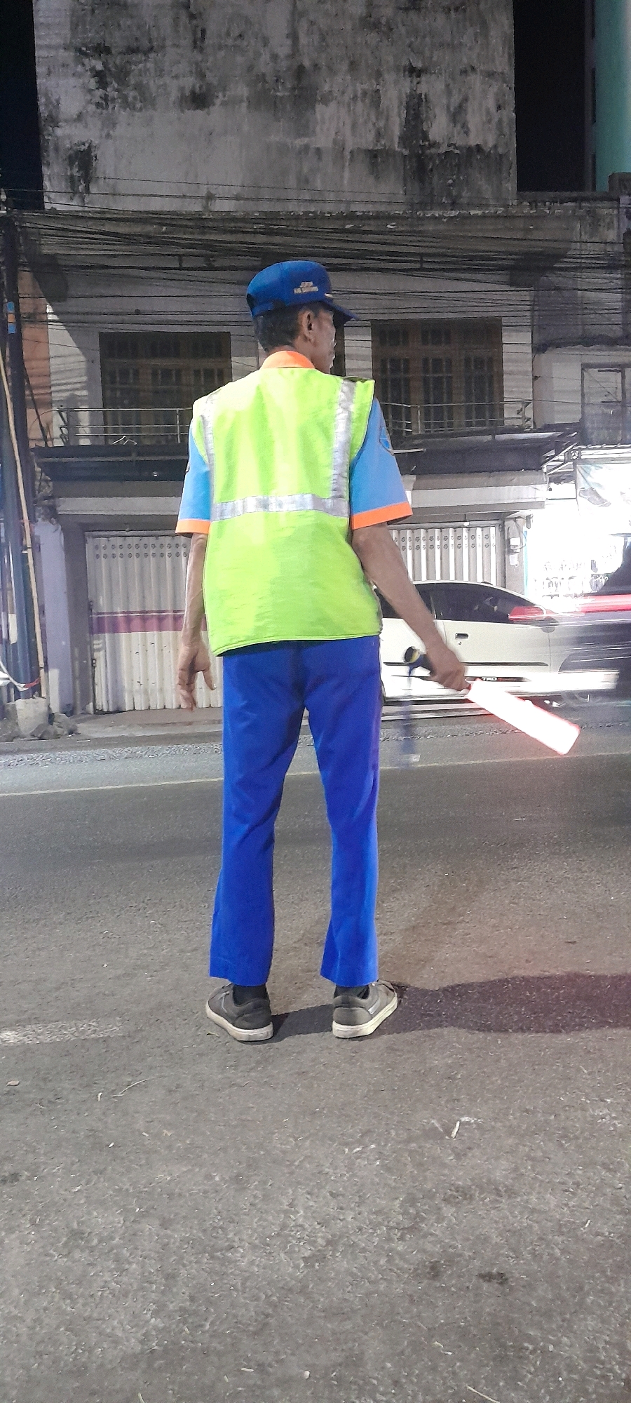 A man wearing a blue cap, a bright yellow reflective vest over a blue and orange short-sleeved shirt, and blue trousers stands on an asphalt road, facing away from the camera. He is holding a red and white luminous baton in his right hand. The background shows a weathered building with multiple windows and what appears to be a garage door. Power lines are visible above the building. A light-colored car is partially visible to the right, and its red brake lights are reflected on the road. The scene appears to be in a city at night, possibly in Rogojampi, Indonesia.