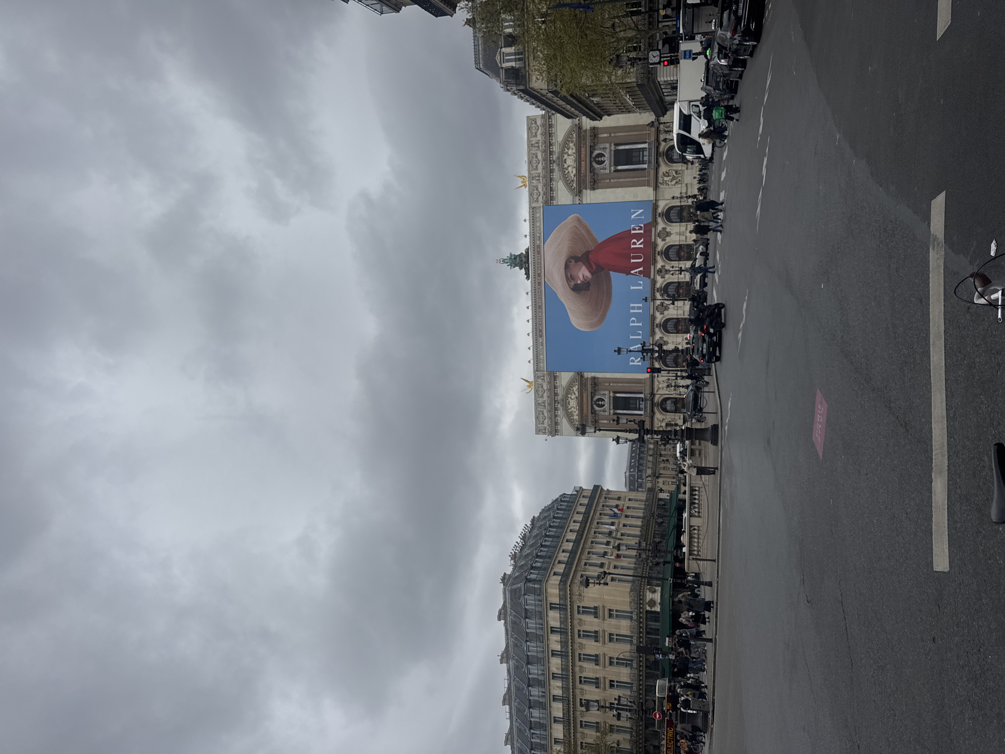 The image is taken from a street-level perspective in Paris, France. The sky is overcast with heavy clouds. In the center, a large advertisement for Ralph Lauren is displayed on the side of a grand building, likely the Palais Garnier opera house. The ad features a woman wearing a large hat. The street is lined with parked vehicles, including cars and motorcycles, and pedestrians are visible walking along the sidewalks. The architectural style of the buildings is consistent with traditional Parisian structures. The street appears to be wet, possibly from recent rain.