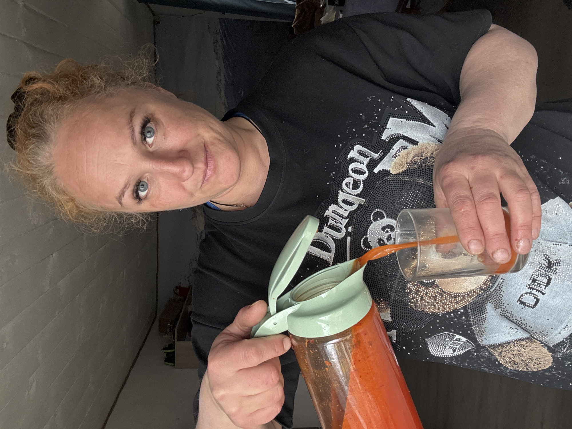 An indoor, eye-level portrait shows a woman in Chaikovsky, Russia, actively pouring a vibrant orange-red liquid from a clear glass pitcher into a clear drinking glass. She holds the pitcher with her left hand, with her thumb on the light green lid's opening mechanism, and supports the drinking glass with her right hand.
The woman appears middle-aged with fair skin, striking blue eyes, and light, curly hair pulled back. She looks directly at the camera with a neutral to slightly pleasant expression. She is wearing a black t-shirt embellished with sparkly silver and gold designs. Visible text on her shirt includes the word "Dungeon" and a partial word that appears to be "Dear" near the bottom right. A design resembling a panda bear is also present on her shirt.
The background consists of a light-colored wall, possibly painted brick or horizontal paneling, on the left. To the right and behind her, the scene recedes into a darker area with indistinct elements, suggesting a corner or passageway within a room. The lighting indicates an indoor setting, possibly with artificial light or diffuse daylight. The overall atmosphere is casual and domestic.