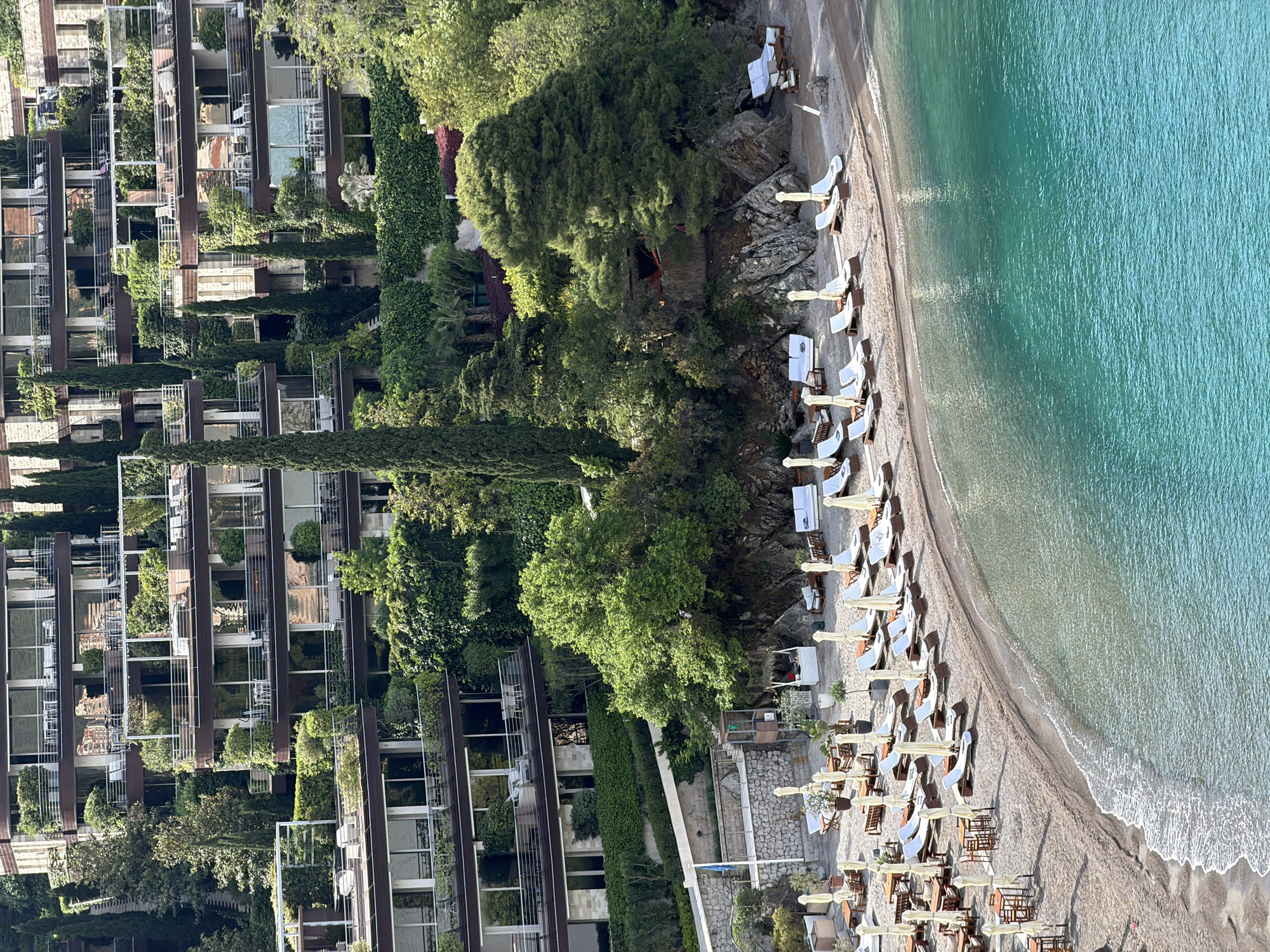 This is an aerial view of a beachfront scene in Budva, Montenegro. A tall, modern building stands to the left, its facade lined with greenery. Lush trees and vegetation fill the space between the building and the beach. The beach itself is narrow, stretching along the coast with numerous lounge chairs and umbrellas set up. The clear turquoise water of the Adriatic Sea extends to the right, showing visible wave action. The scene appears calm and sunny. There are no people visible on the beach.