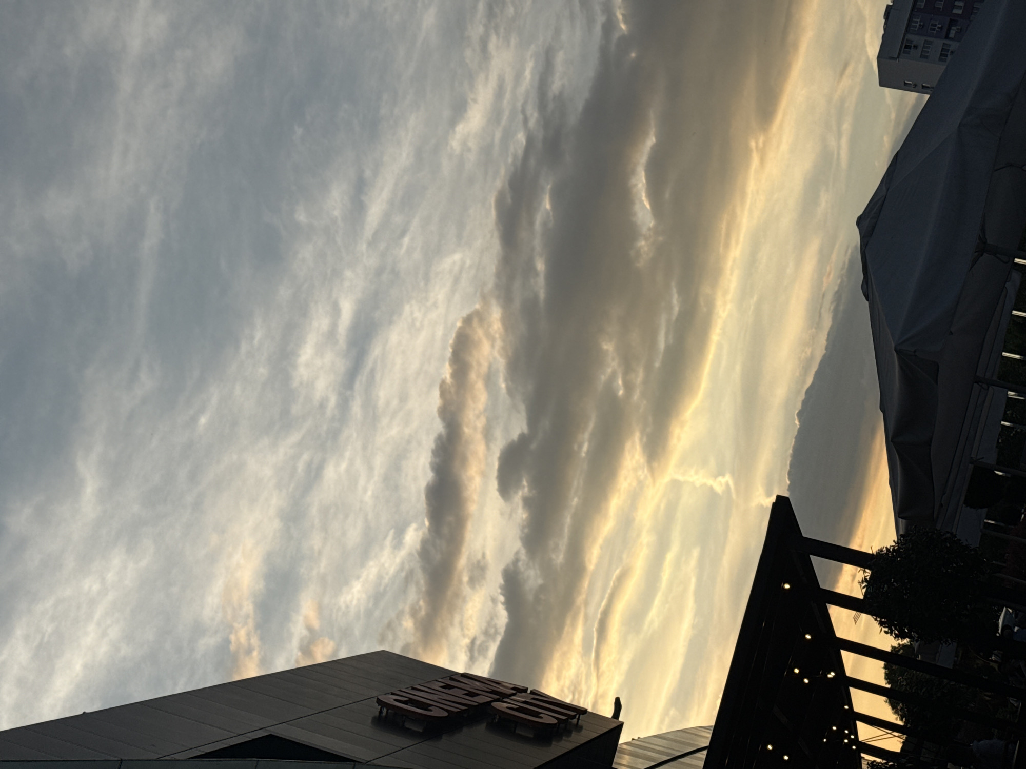 The image captures a sky with varied cloud formations, ranging from thin, wispy cirrus clouds to thicker, more defined altocumulus or stratocumulus clouds. The lighting suggests a sunset or sunrise, with golden hues illuminating parts of the clouds.
In the lower portion of the frame, architectural elements are visible. A dark, modern building with a prominent sign that says "CHIMES" stands out. Further in the background, there is a glimpse of a taller, residential or commercial building. The presence of a canopy and other elements like trees suggest the scene is in an outdoor setting, possibly a patio or terrace.
Based on the sign "CHIMES," this could be a restaurant or bar, located in Burgas, Bulgaria. The building with the Chimes sign is in front of a setting with an outdoor tent-like structure and surrounding trees. The presence of architectural structures indicates it is an urban environment. There are no visible people in this frame.