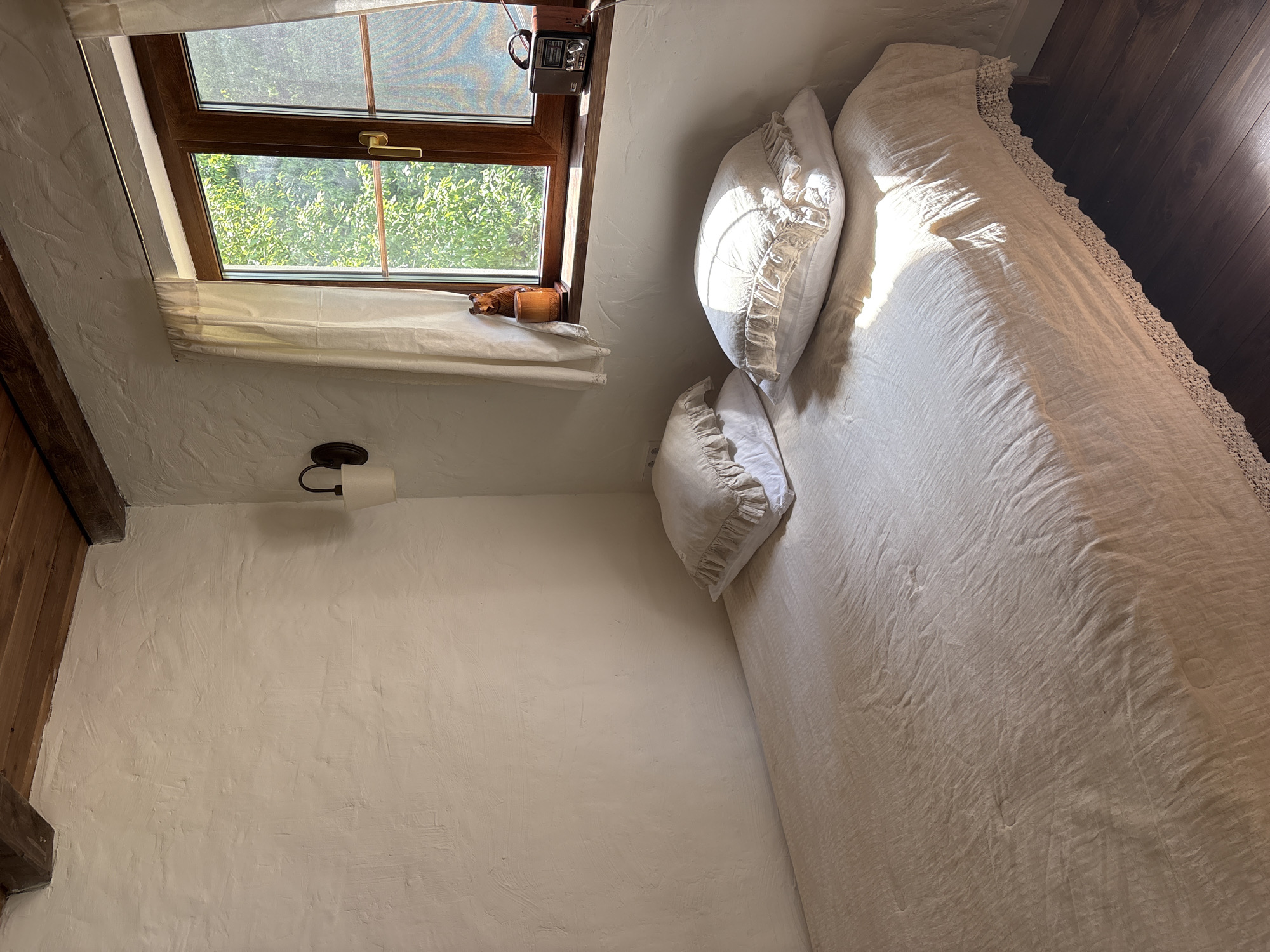 A room with white, textured walls features a wooden-framed window showing greenery outside. A small white curtain covers part of the window. A wooden carved animal figurine and a vintage radio sit on the windowsill. A bed with white linens and decorative pillows occupies a portion of the room. The wooden floor is visible in the upper right. A dark-colored light fixture is attached to the wall. The location appears to be inside an interior space, potentially a bedroom. It is not possible to confirm the location is in Sibir', Russia, from the visual information provided.
