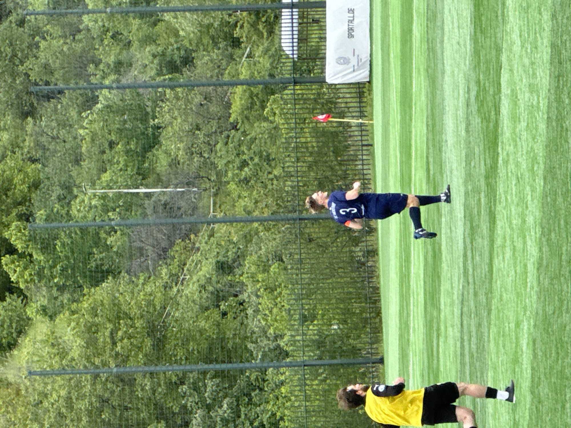 The image shows two soccer players on a green field, potentially an artificial surface, with surrounding trees. One player is wearing a dark blue uniform with the number "3", while the other wears a yellow jersey and black shorts. A banner or sign with "SPORTALL.GE" is visible. A fence separates the playing field from the tree line. The scene suggests a match or practice in Tbilisi, Georgia.