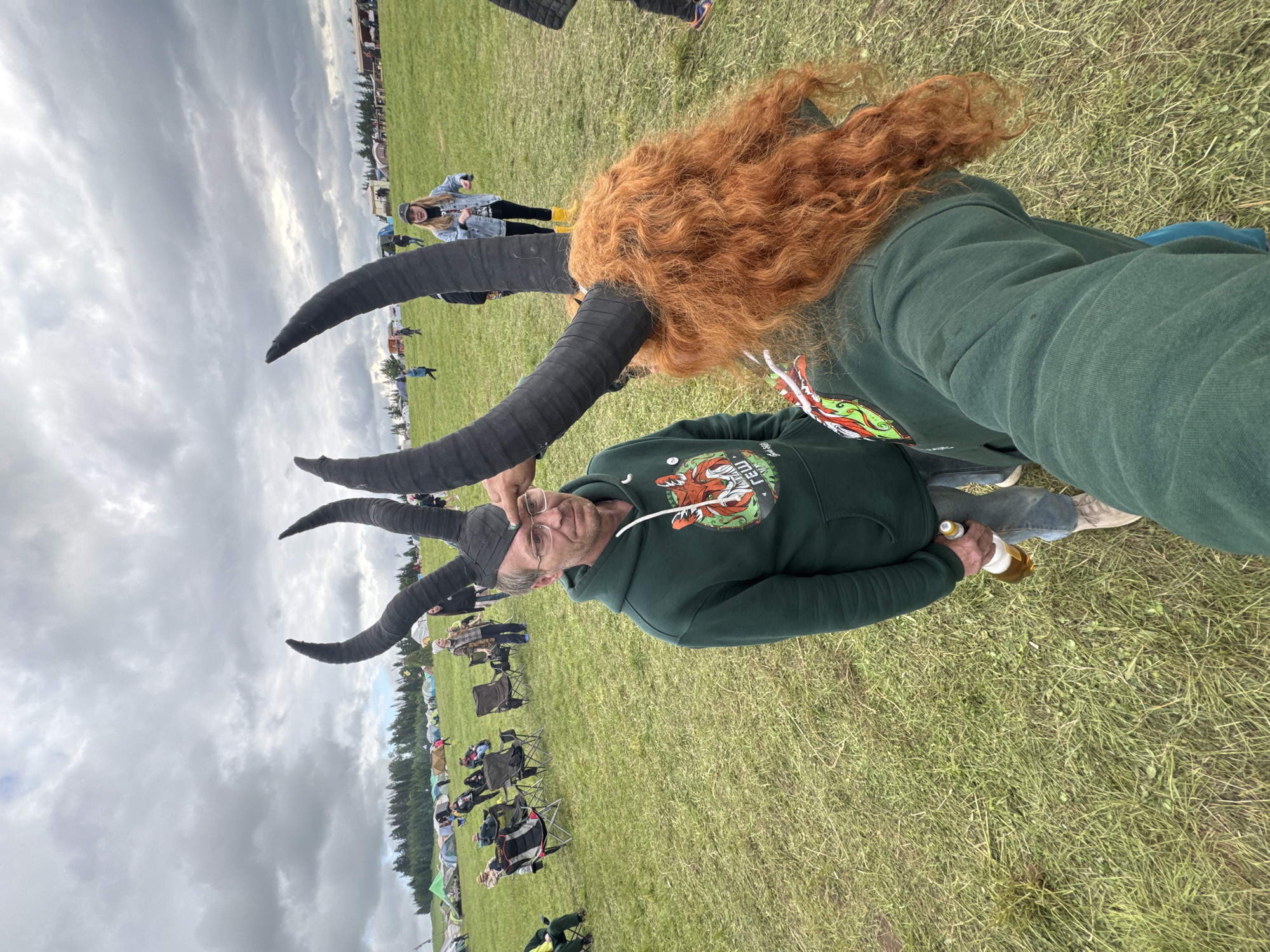An outdoor scene captures a large gathering on a grassy field under a heavily overcast sky in Большая Кивара, Russia. In the immediate foreground, a person with long, curly, reddish-brown hair is seen from behind, wearing a dark green hoodie with a colorful emblem on the left shoulder and a black headpiece featuring two large, upward-curving black horns. Facing this individual, to the left of the frame, stands a Caucasian male with short, light brown hair and glasses. He wears a matching dark green hoodie with an identical colorful emblem depicting a fox-like creature and the text "ГЕШ," blue jeans, and light-colored shoes. He holds a clear glass bottle containing a yellow liquid in his left hand. His right hand is raised, appearing to touch or adjust one of the black horns on the headpiece of the person taking the photograph. His facial expression is neutral. In the mid-ground and background, numerous camping tents of various colors (green, blue, white, and red) are scattered across the field, indicating a large encampment. Multiple individuals are visible throughout the background, some seated in portable camping chairs, others standing or walking. A young woman in the mid-ground right wears a denim jacket, a baseball cap, and prominent yellow boots. A dense line of evergreen trees forms the distant horizon, beneath a vast expanse of gray clouds. The ground is covered in green grass with some drier patches.