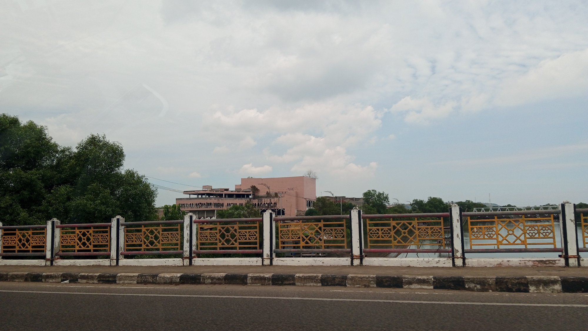 The image shows a view from a bridge over a body of water, with a distinct pink building in the background. The foreground features a road with a concrete barrier and a decorative metal fence along the edge of the bridge. The fence has alternating yellow and red horizontal bars with white pillars. The background reveals lush green trees and the aforementioned pink building, which appears to be under construction or in a state of disrepair, with visible scaffolding and unfinished sections. The sky is cloudy, with patches of blue peeking through. The presence of the pink building, known locally as the Lhokseumawe City Hall or Civic Center, indicates this is likely a location within Lhokseumawe, Indonesia. There are no people or animals visible in the frame.