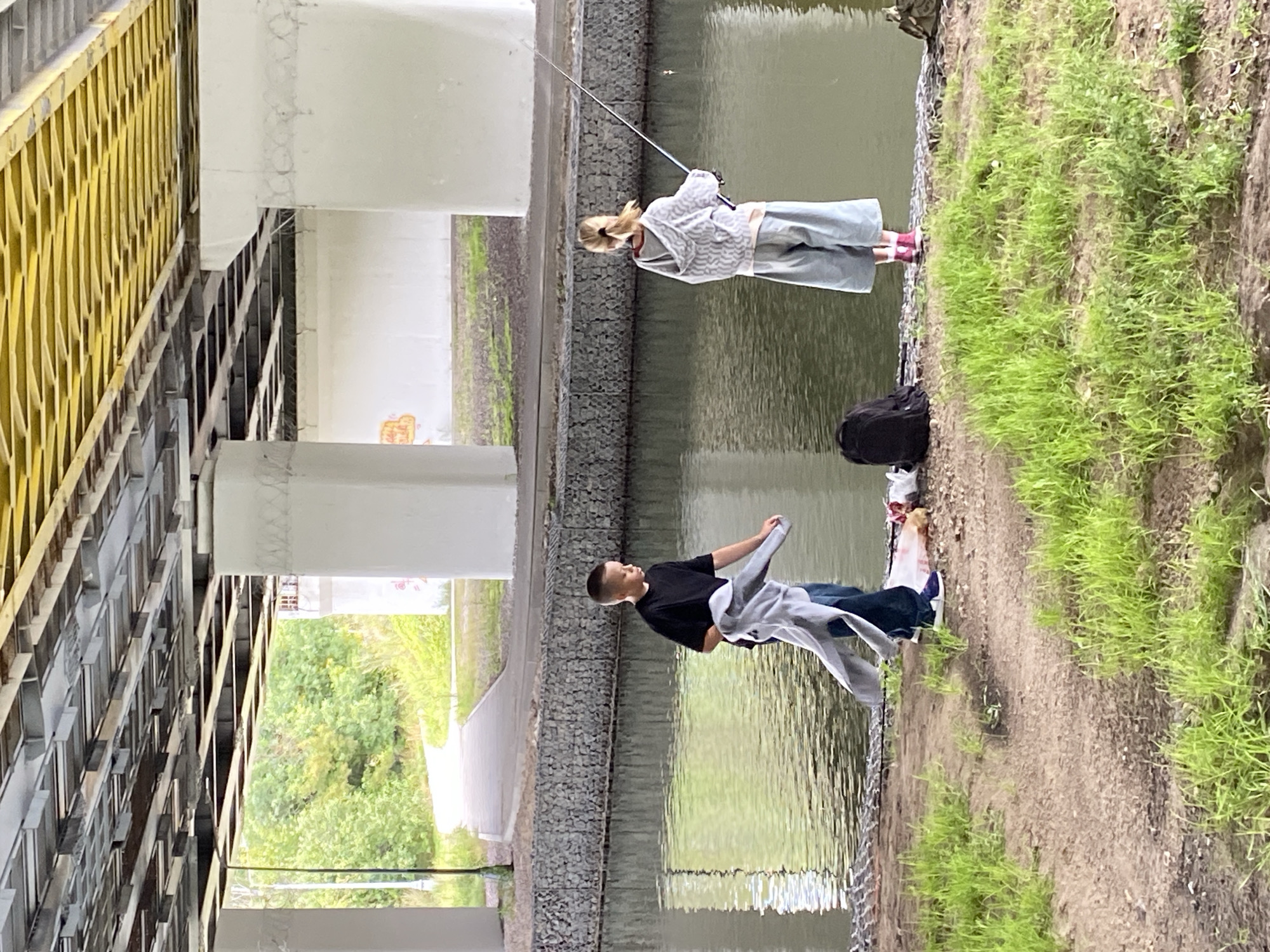 Underneath a bridge, two children are fishing by a body of water. The boy, on the left, wears a black t-shirt and jeans, holding a gray garment. The girl, on the right, has blonde hair tied back in a ponytail and wears a gray hooded jacket and wide-leg pants, holding a fishing rod. A black backpack is visible on the bank behind her, and a pile of items, including what appears to be fish, lies on the ground near the boy. The ground is a mix of dirt and grass, and the water is calm. In the background, a paved path winds through green foliage under a bright sky, with concrete pillars supporting the bridge structure.