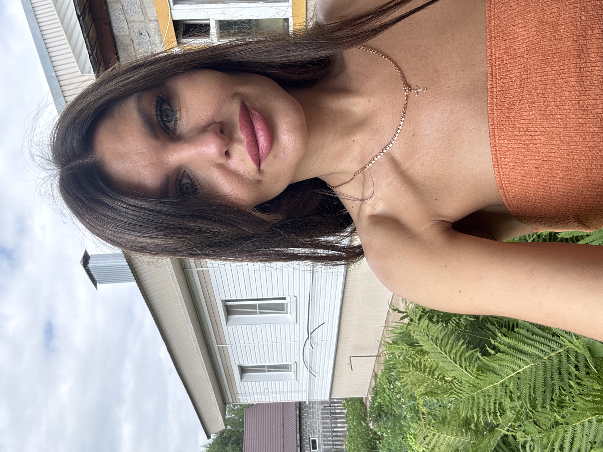 A woman with dark brown hair and light blue eyes, wearing an orange strapless top and a gold necklace with a cross pendant, is captured in a selfie outdoors. She is positioned in the foreground, with a white house and lush green ferns forming the background. The house has a light beige foundation and two white-paneled windows. A silver chimney with horizontal ridges is visible on the roof. A darker-colored roof and a wooden fence are also present in the background. The sky above is overcast with scattered clouds.