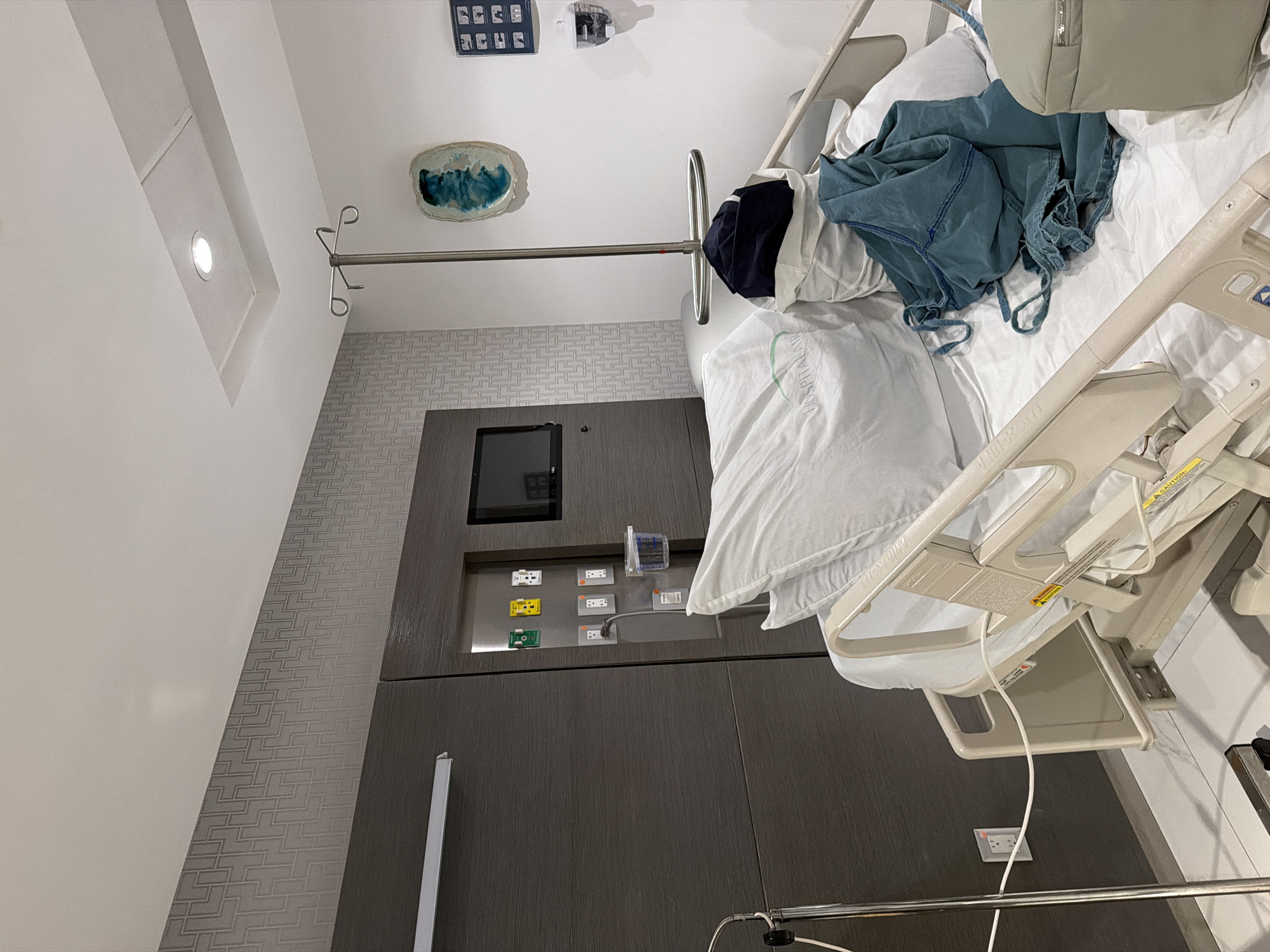 The image shows the interior of what appears to be a hospital room. A hospital bed occupies the right side of the frame, with white sheets, a pillow, a blue garment, and a beige bag on top. An adjustable metal IV stand extends towards the center of the image. The left side of the frame shows a wall with a light fixture in a recessed panel, a textured wallpaper section, and a gray built-in unit with a monitor and a panel of electrical outlets. A piece of art adorns the wall above the unit. The scene suggests a medical or healthcare setting. The location within Villahermosa, Mexico, is not determinable from the image.