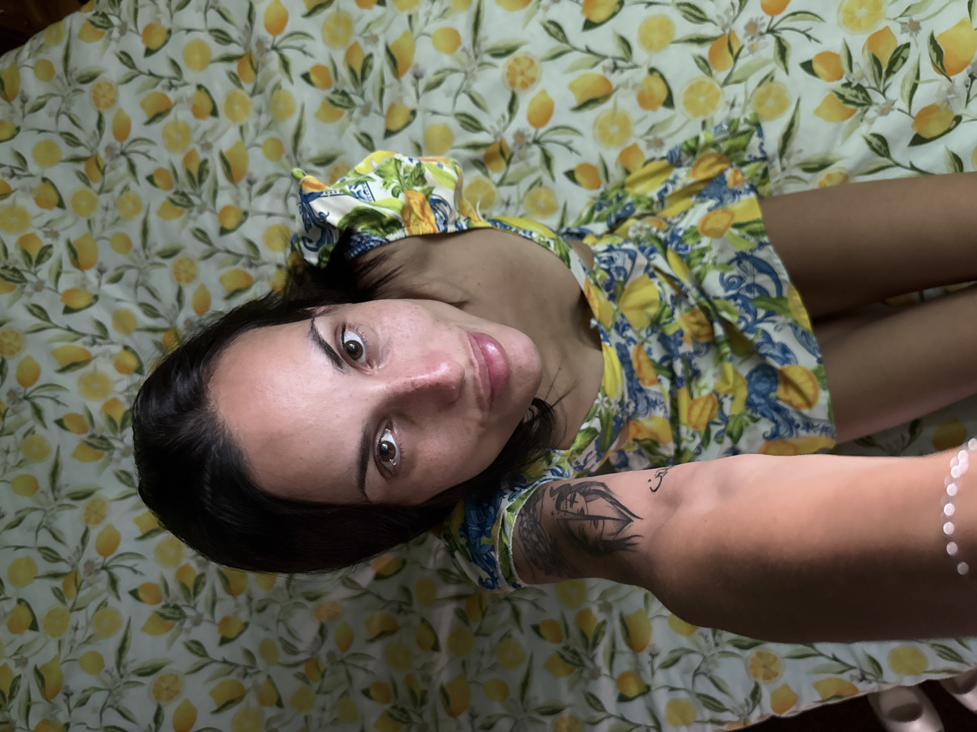 A woman lies on a lemon-patterned sheet, looking directly at the camera. She wears a blue and yellow dress with a similar lemon print. The woman has dark hair and brown eyes. Her left arm, adorned with a tattoo, extends towards the camera. The location is unknown; the scene shows only a bedroom interior and does not include sufficient detail to infer the location in the city of Pleven, Bulgaria.