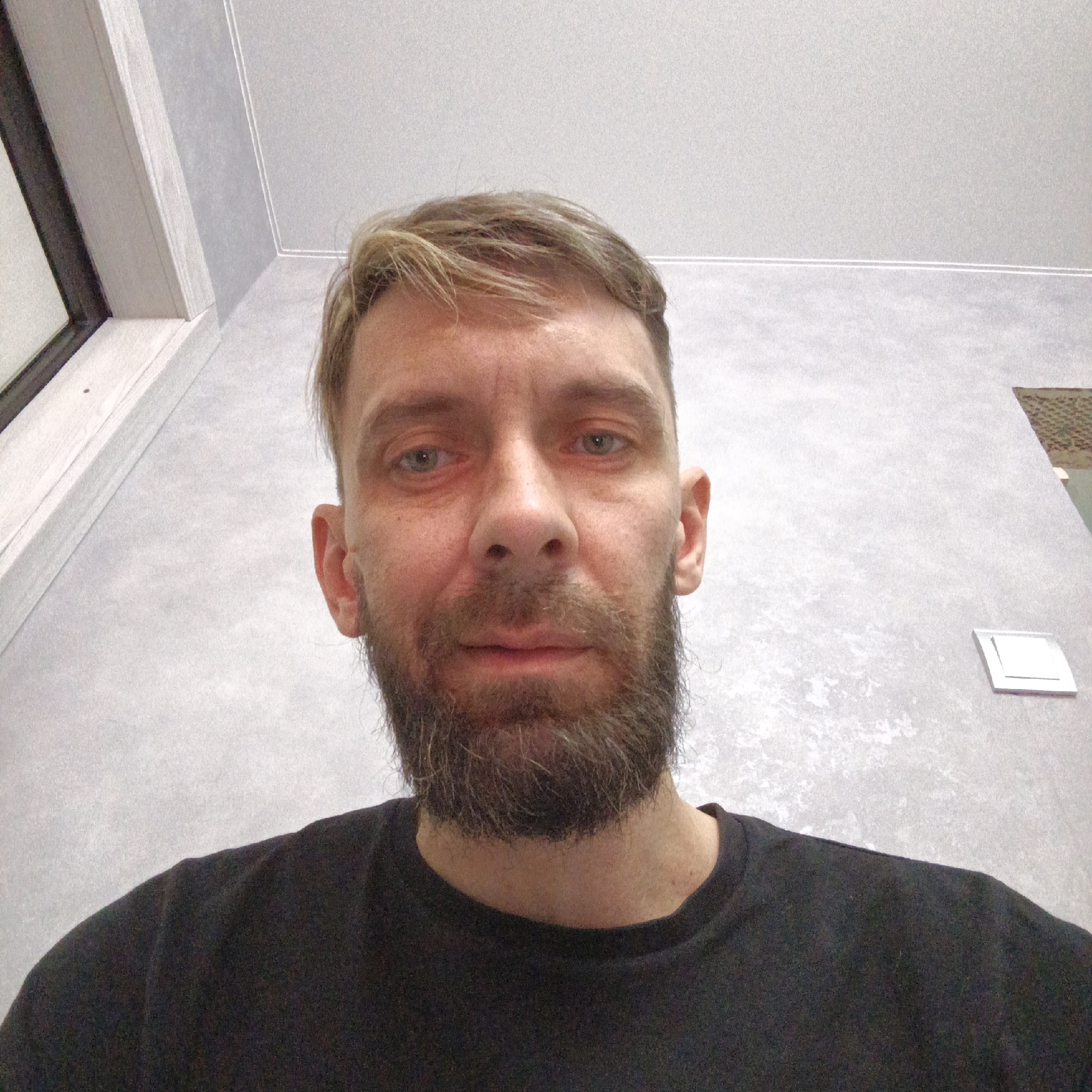A middle-aged man with a beard and blonde hair, wearing a black t-shirt, is looking directly at the camera. The background is a light gray wall with a window and a ventilation grill visible. The image was taken indoors. There is no definitive information suggesting the image's location is in Марковский, Russia.